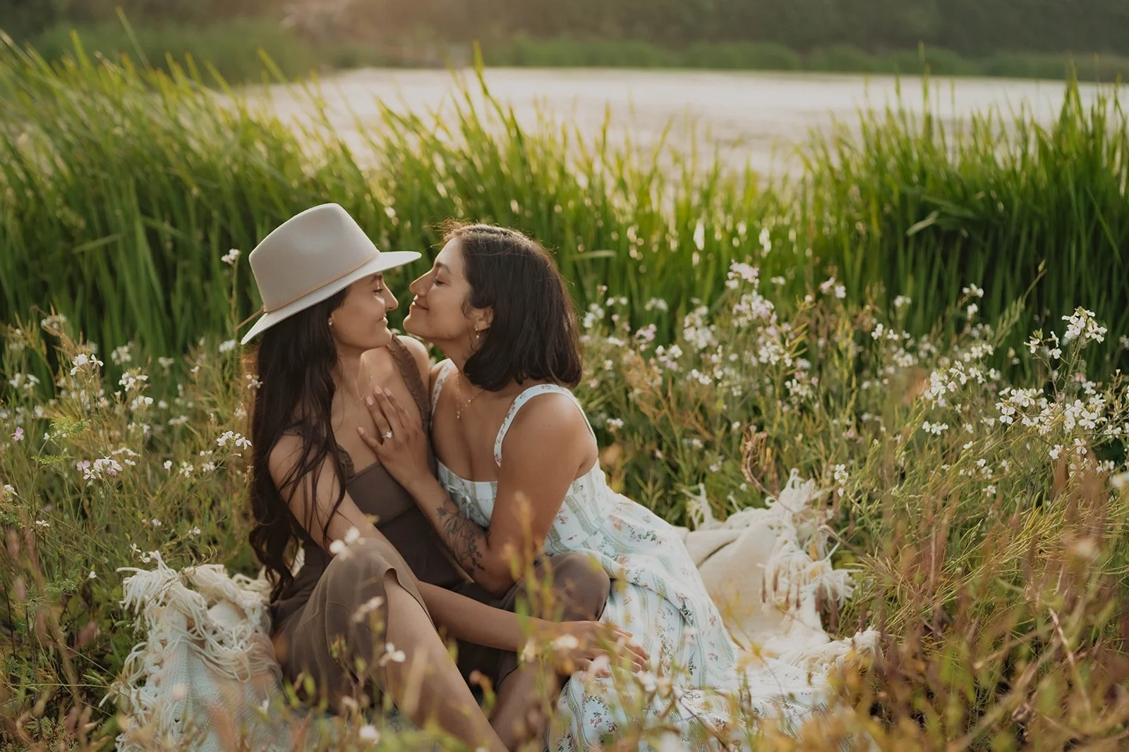 Two women sitting on a blanket among tall grass and wildflowers near a body of water, leaning close and smiling at each other, during sunset.