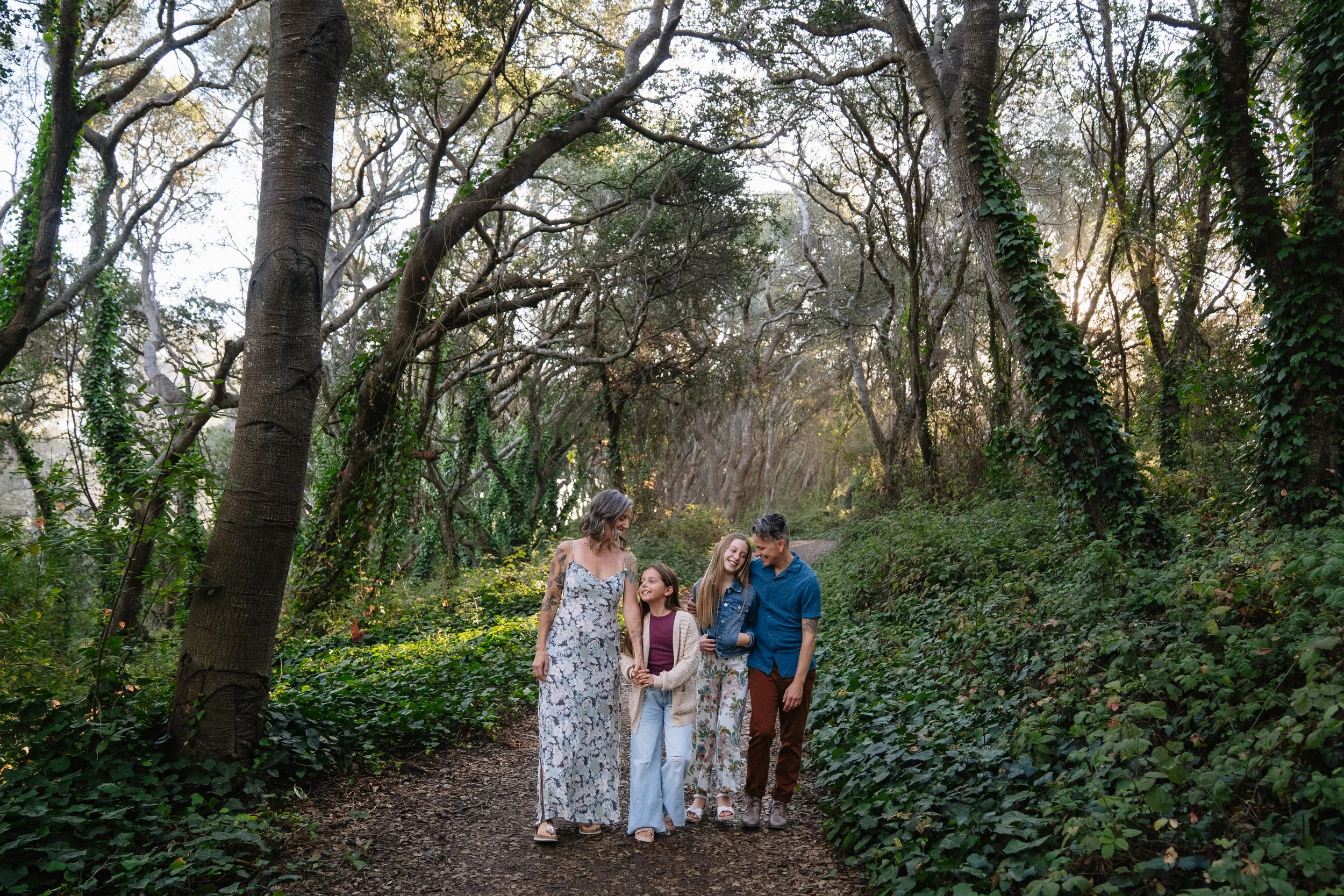 A family of four walking along a forest trail, smiling and talking with each other. The forest has tall trees and green foliage.