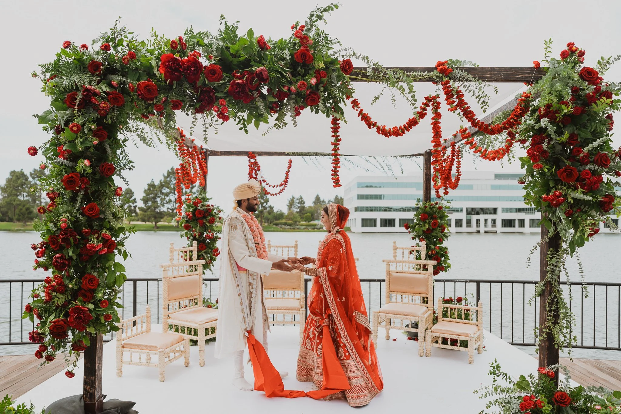 A bride and groom in traditional Indian wedding attire exchanging vows under a decorated floral arch on a lakeside wedding venue.