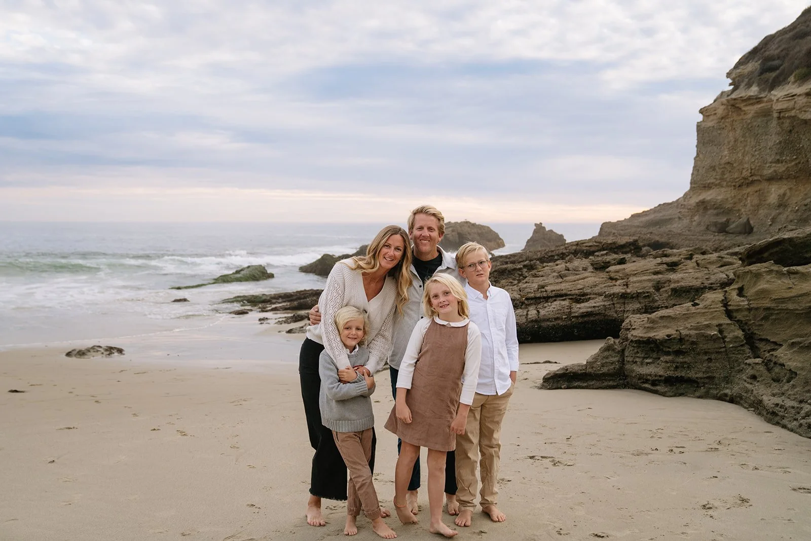 A family of six standing on a sandy beach near rocks and the ocean, smiling for a photo on a cloudy day.
