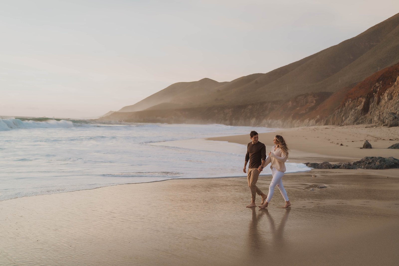 A couple walking hand in hand along the beach near the water, with mountains in the background during sunset.