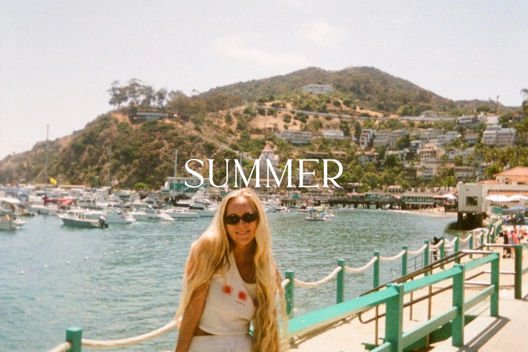 A woman in sunglasses is smiling on a pier by the water with boats, hills, and buildings in the background. The word "SUMMER" is written across the image.