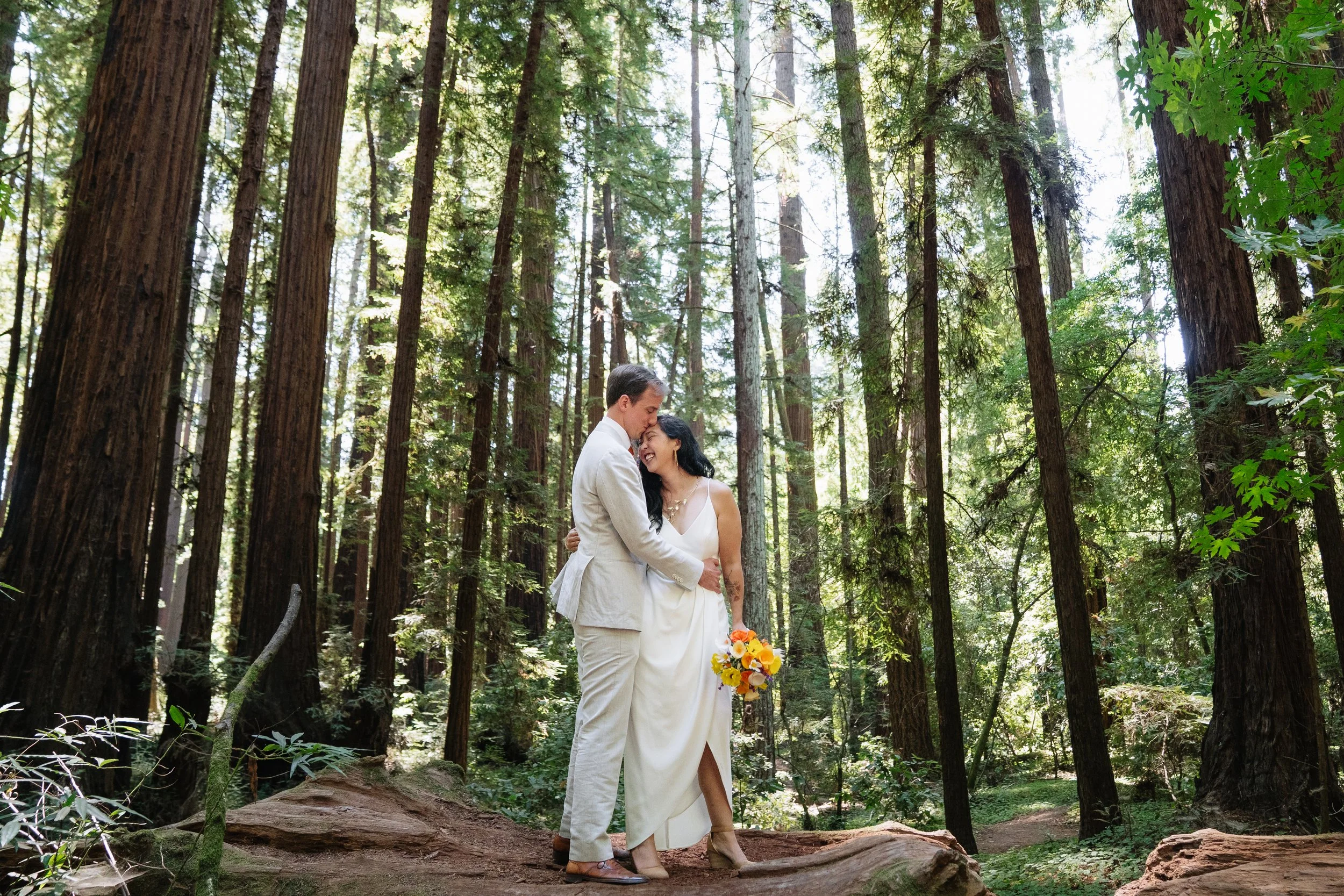 A couple dressed in wedding attire embracing in a forest with tall trees, sunlight filtering through, and the woman holding a bouquet of yellow and orange flowers.