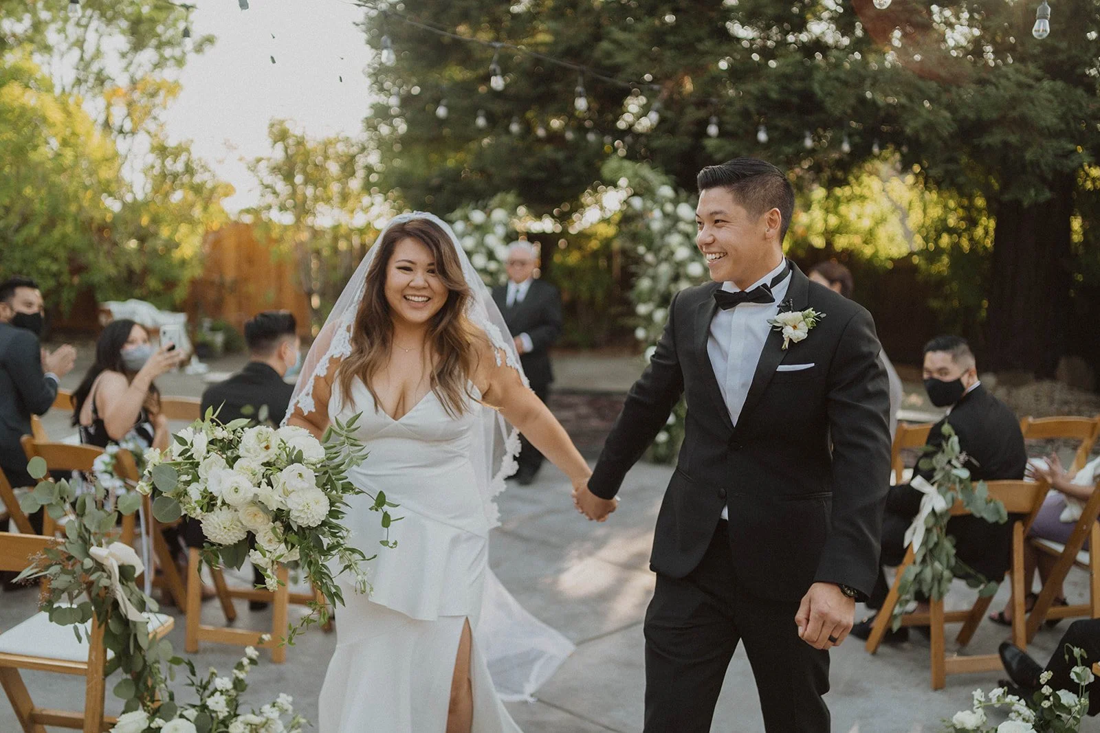 Happy couple holding hands and smiling at their outdoor wedding ceremony with guests seated on wooden chairs decorated with flowers.