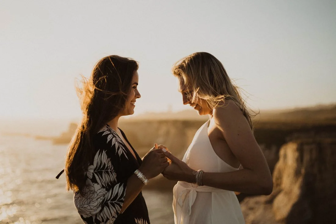 Two women standing close together at sunset by the ocean, holding hands and smiling.