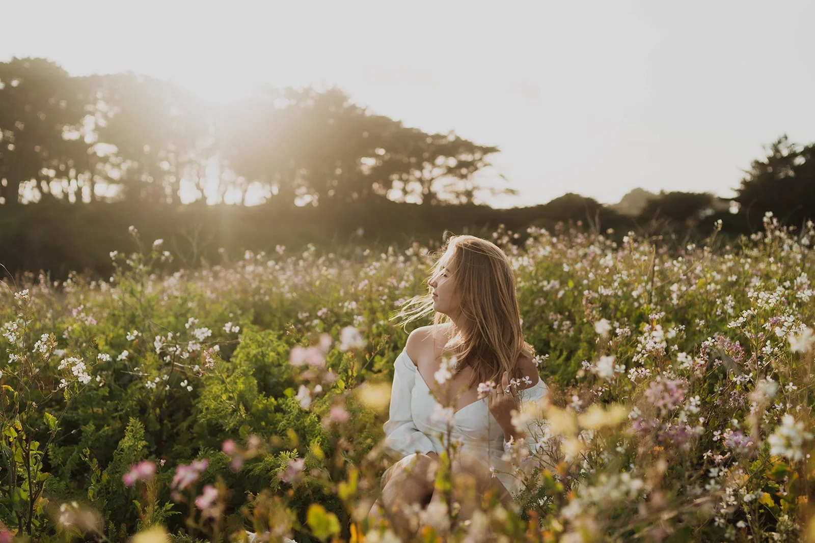 A young woman with long, wavy hair sitting in a field of pink and white wildflowers during sunset, with trees in the background.