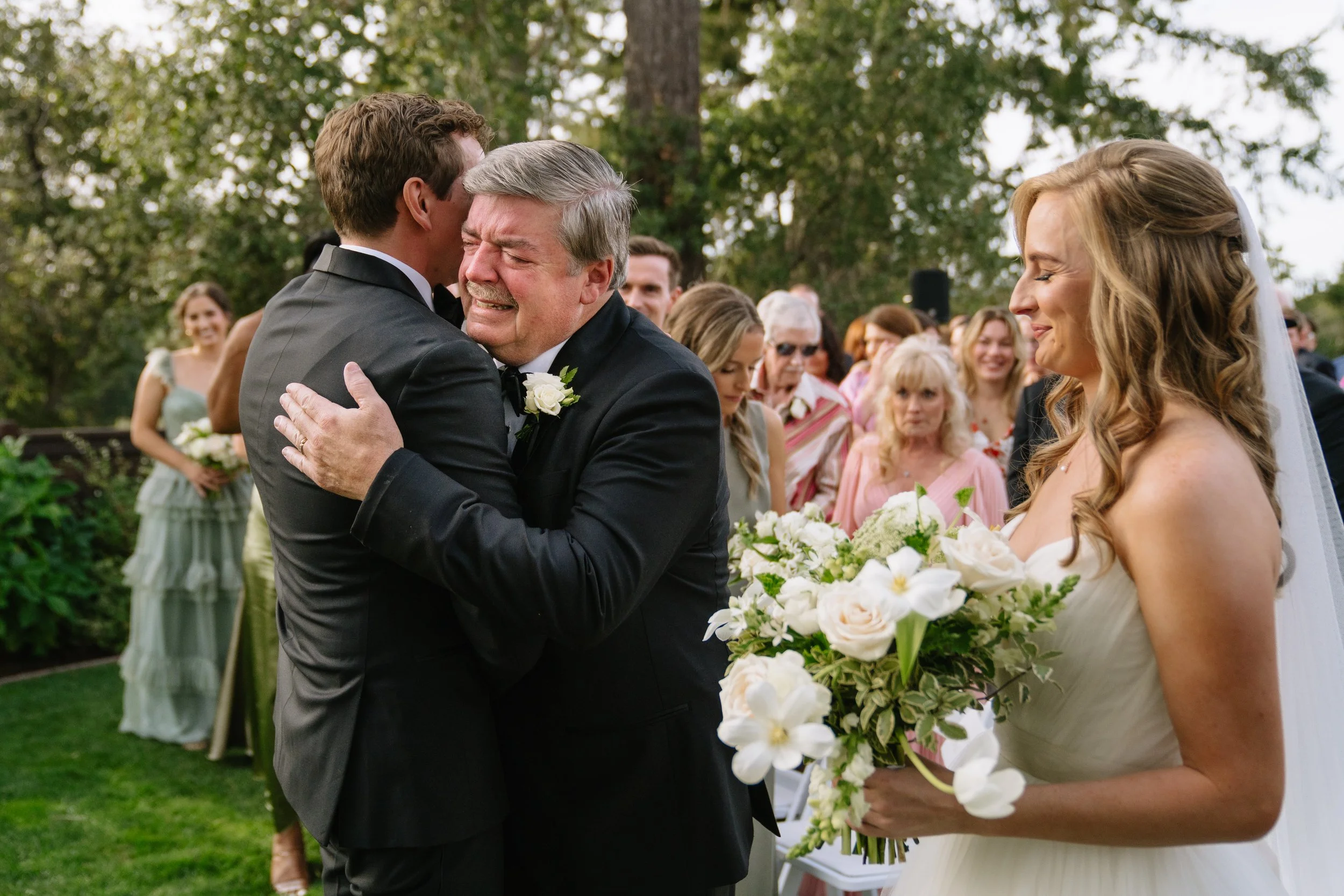 A wedding ceremony outdoors, with a bride holding a bouquet of white flowers, a groom, and a man in a black suit emotional hug. Guests watch in the background, with trees and greenery surrounding the scene.