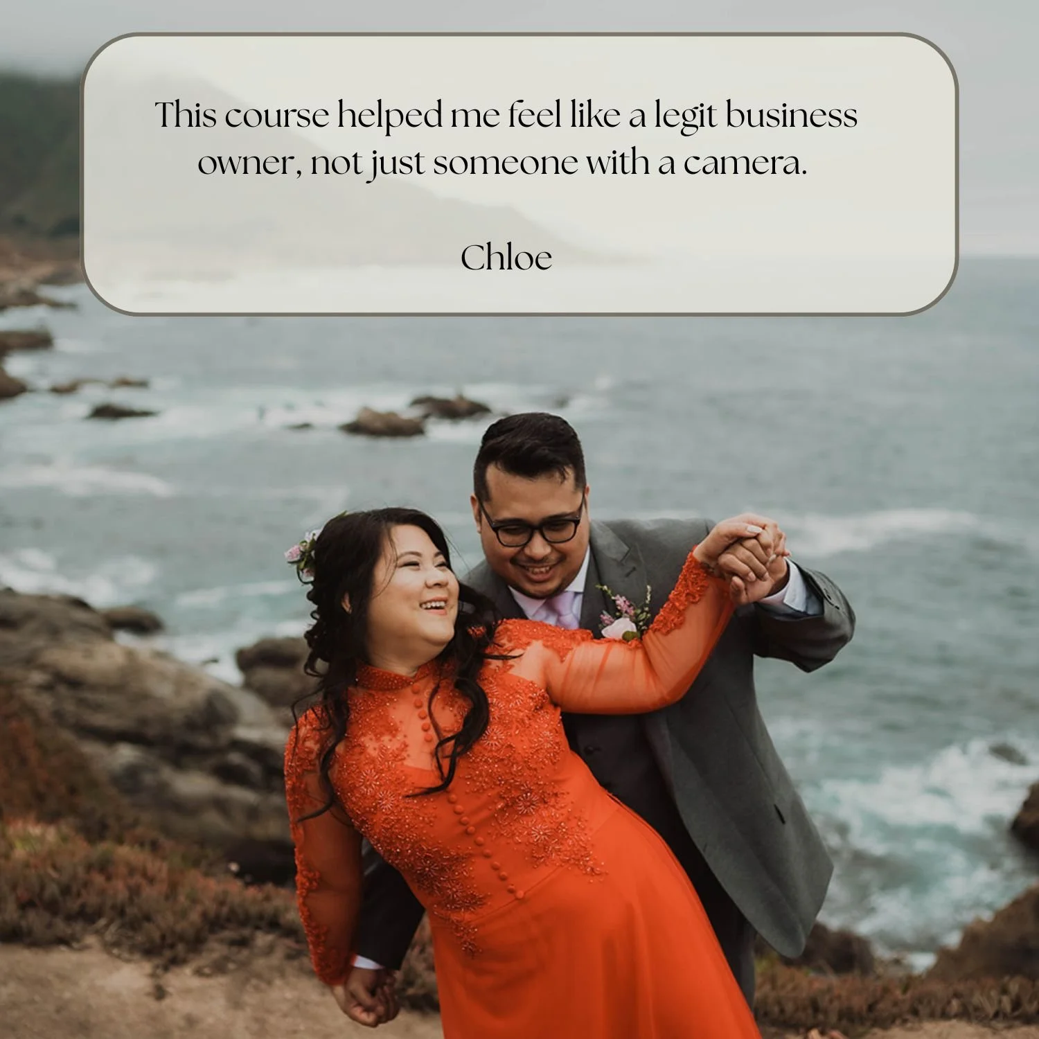 A couple dancing by the rocky coast, the woman in an orange dress and the man in a gray suit, both smiling.