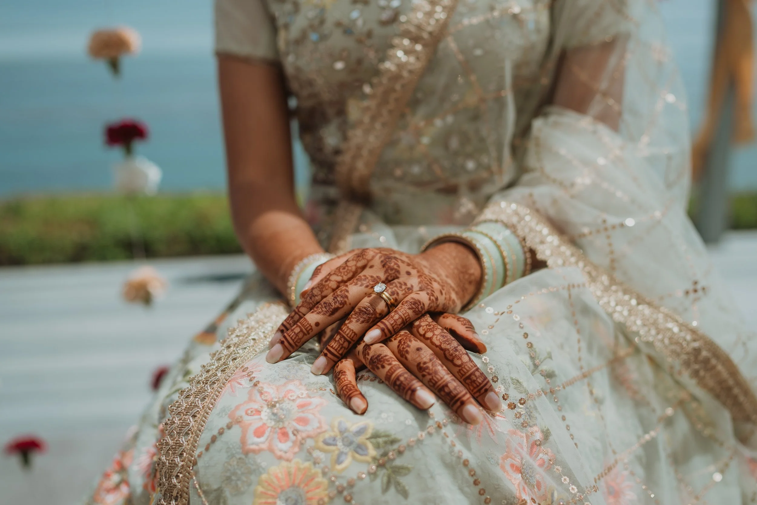 Close-up of a bride's hands resting on her lap, showing intricate henna designs, jewelry, and ring, with a traditional embroidered dress.