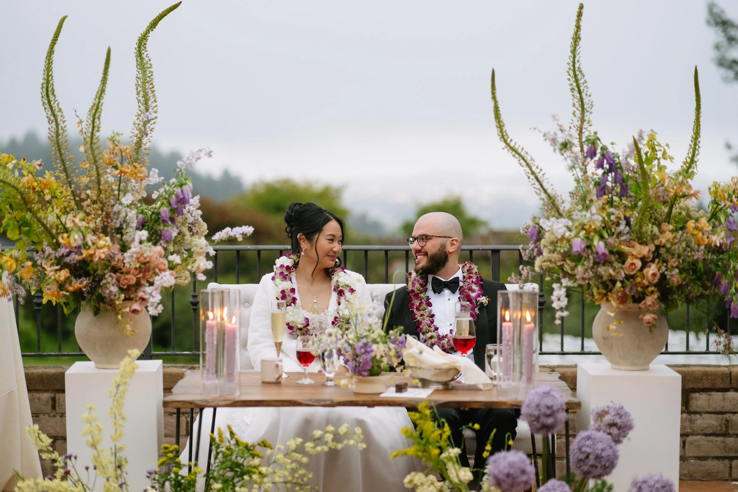 A couple sitting at a decorated outdoor wedding reception table surrounded by large floral arrangements, candles, and glasses of champagne and wine, smiling and looking at each other.