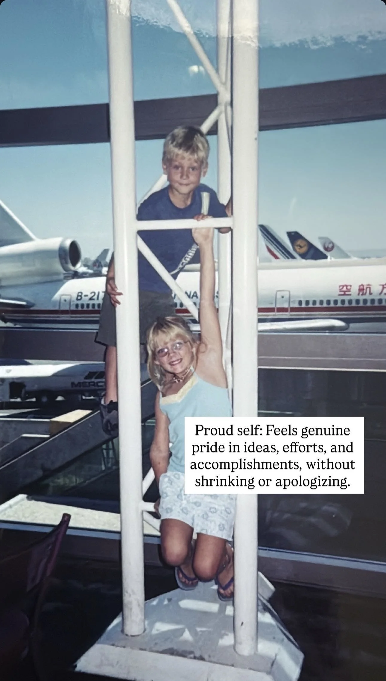A young boy standing on a ladder and holding a young girl who is hanging from the ladder, with airplanes visible in the background at an airport.