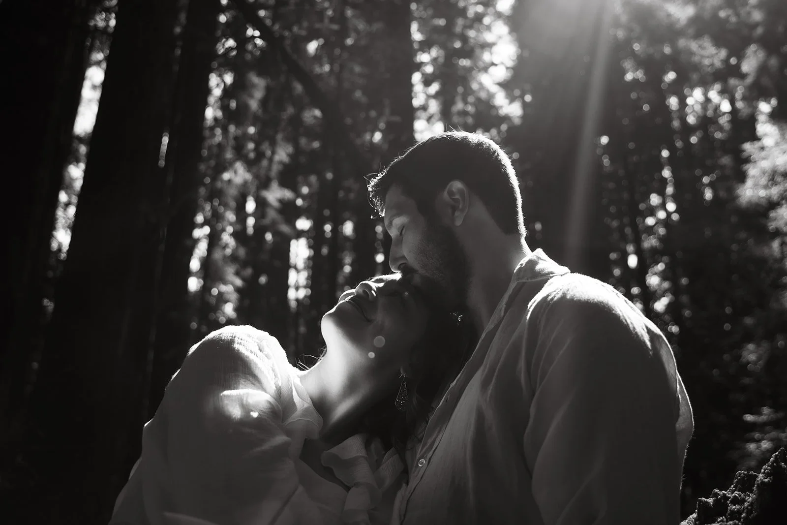 A black and white photo of a couple in a forest, sharing a kiss with the sunlight shining through the trees.
