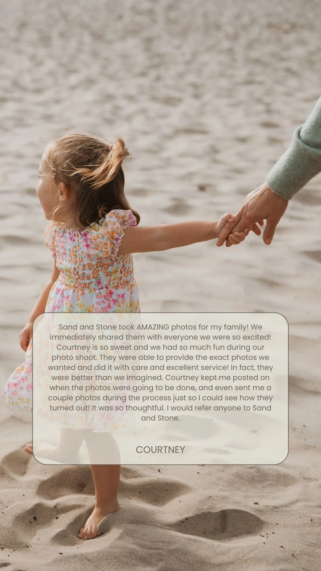 A young girl in a floral dress holding hands with an adult on a sandy beach with water in the background.