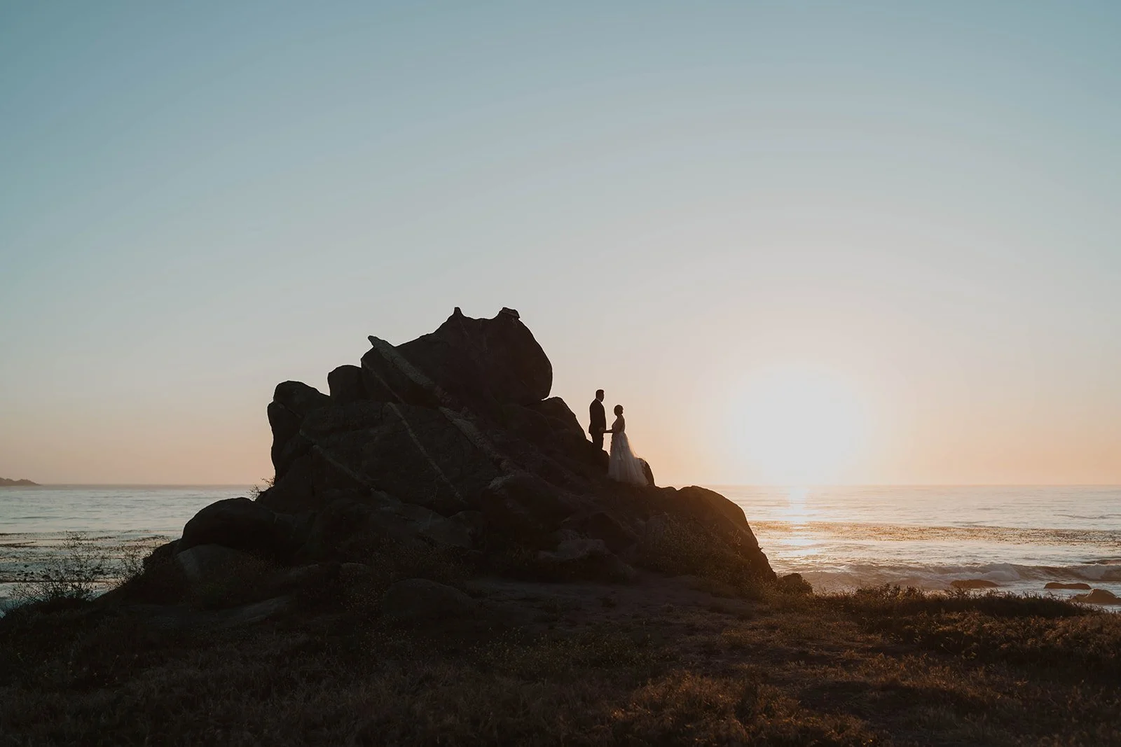 Silhouette of a couple standing on a rocky outcrop by the ocean during sunset.