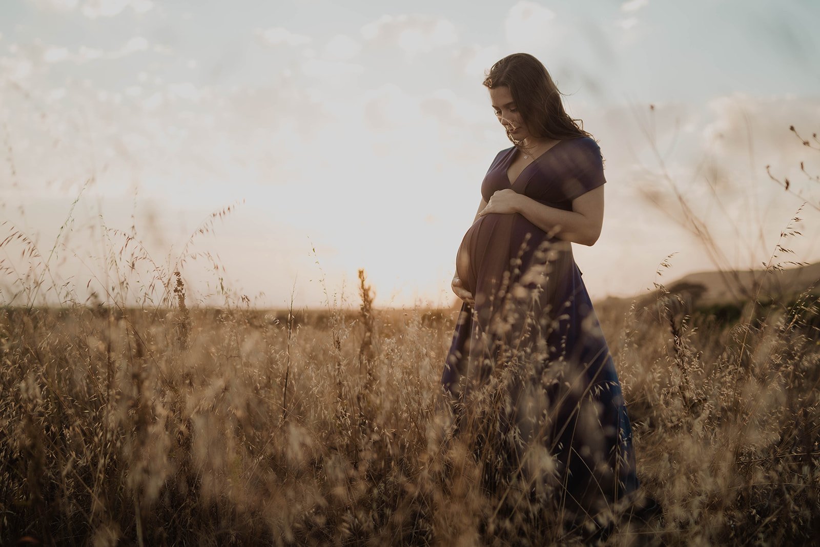 Pregnant woman standing in a field of tall grass during sunset, holding her belly with both hands, wearing a dark dress.