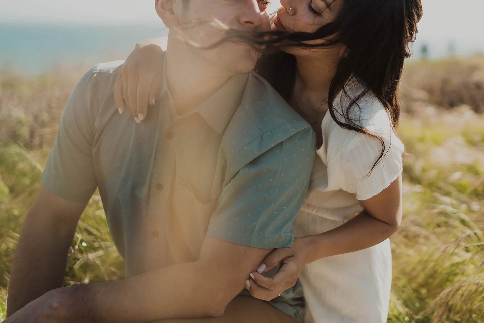 A couple sharing a kiss outdoors on a sunny day, with the man leaning towards the woman and the woman holding onto the man's arm. The background is blurred with greenery and a bright sky.