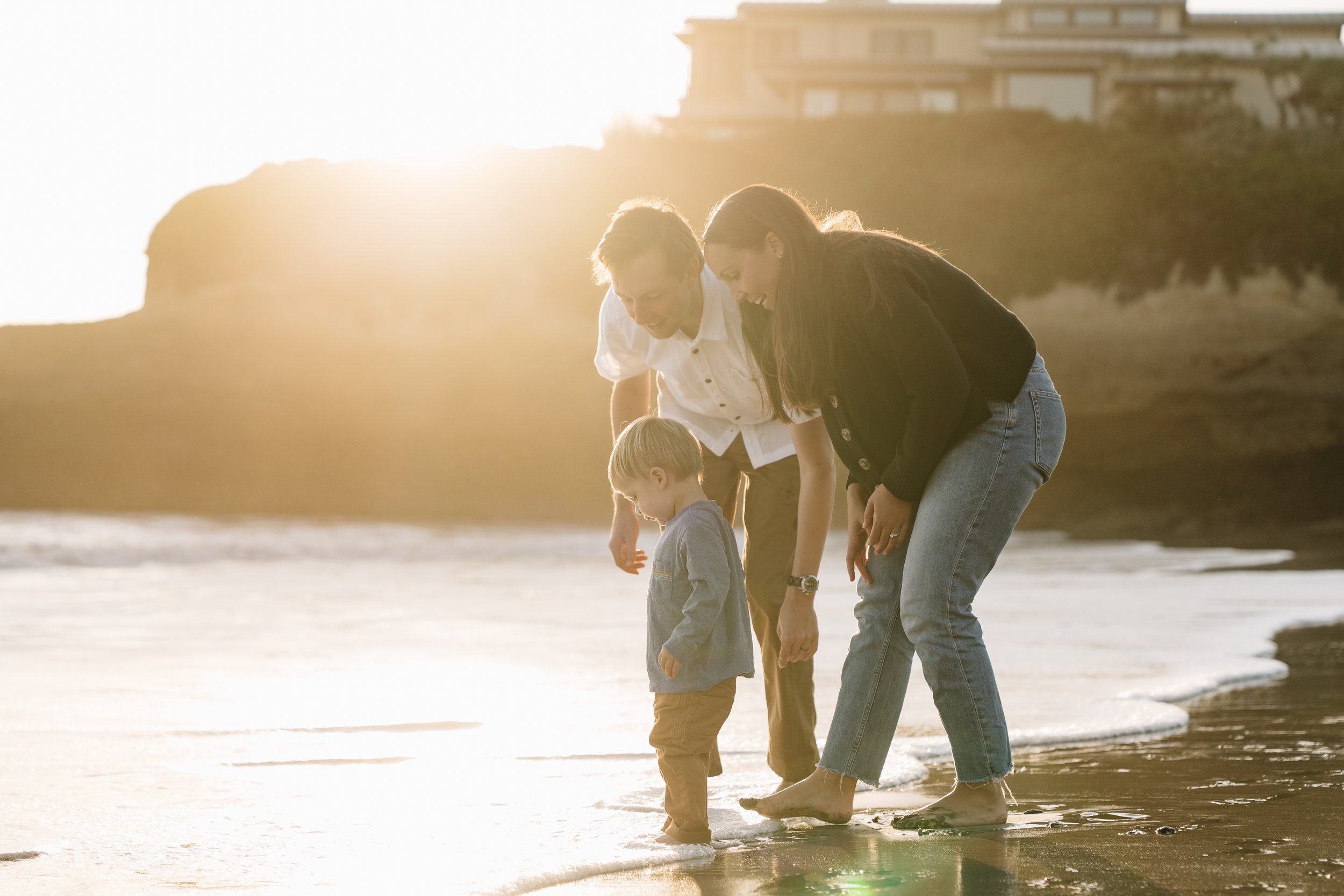 Family of three, two adults and a young boy, walking barefoot along a beach at sunset.