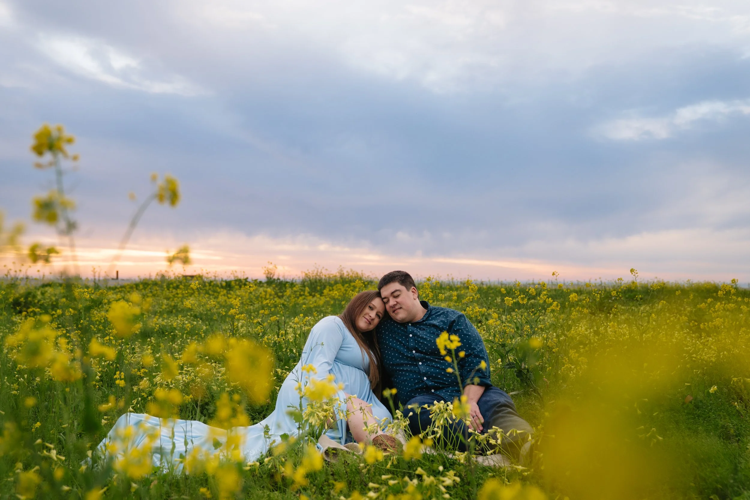 A couple sitting in a yellow flower field at sunset, with cloudy sky overhead.