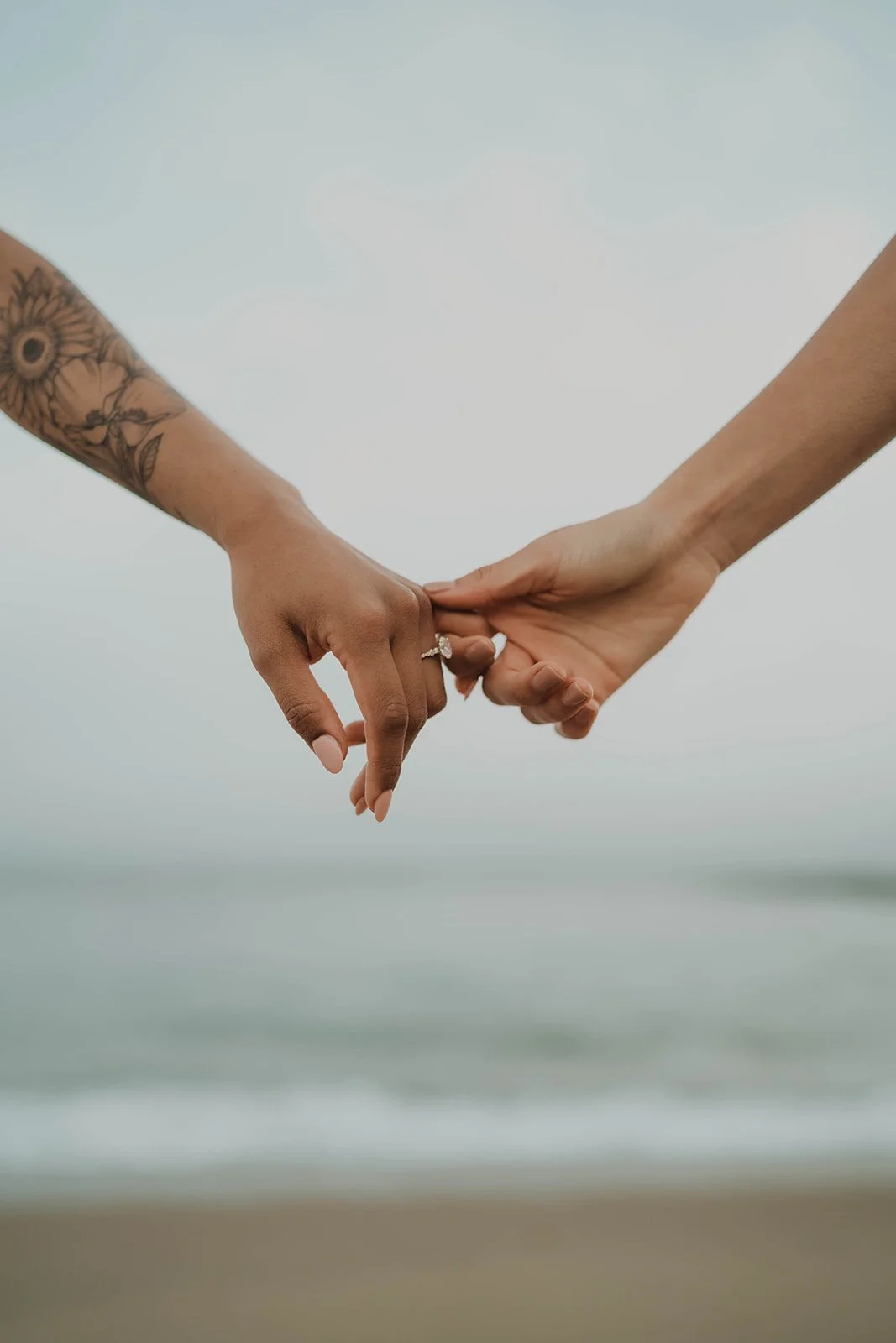 Two people holding hands with their fingers intertwined, one has a floral tattoo on the arm and an engagement ring, the background is a blurred beach scene.