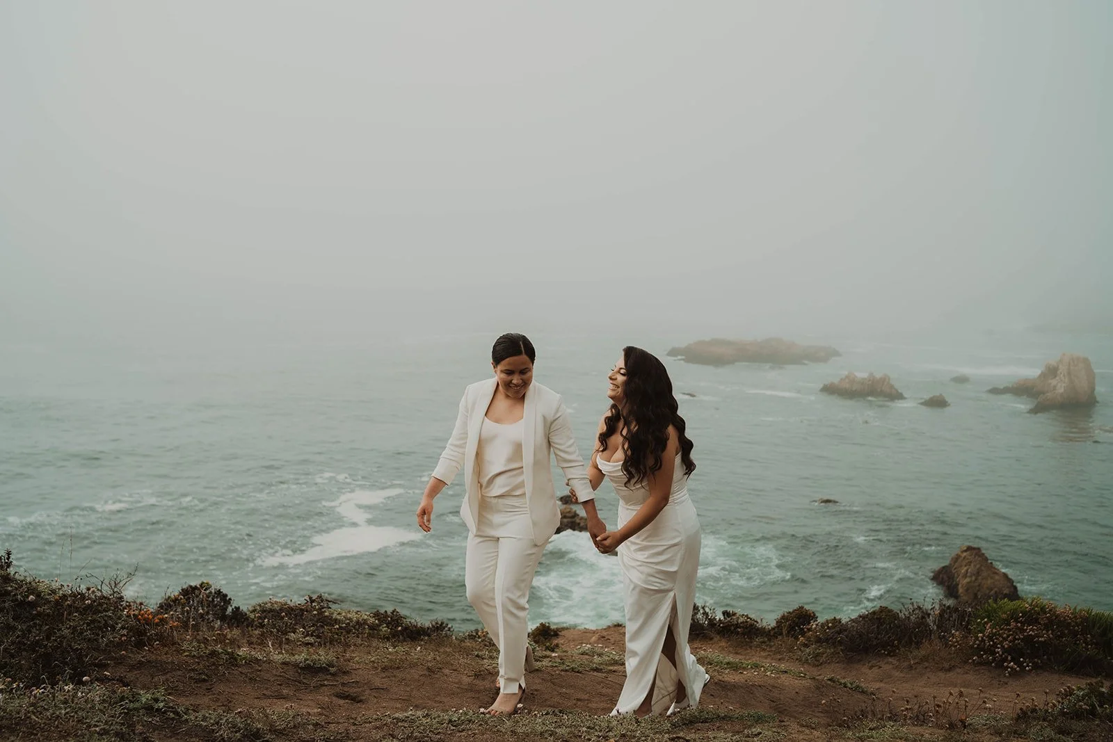 Two women walk hand-in-hand along the coast, dressed in white, with the ocean and rocks in the background, amidst foggy weather.
