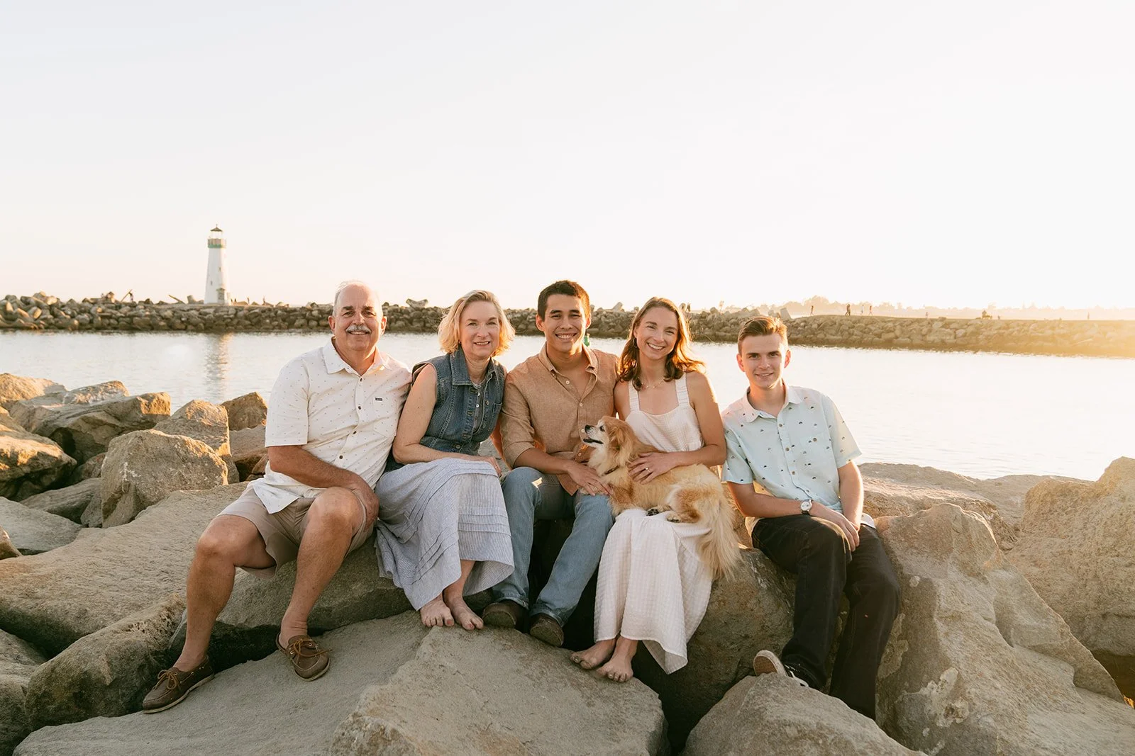 A family of five with a dog sitting on rocks near water with a lighthouse in the background during sunset.