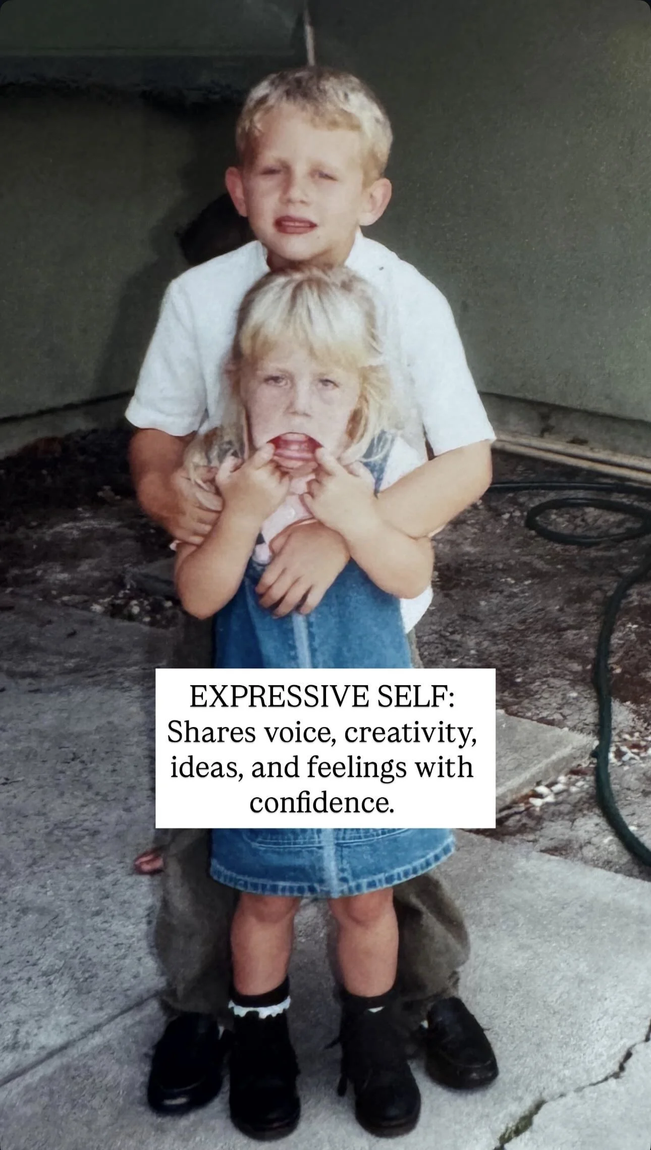 A young boy with blond hair and a serious expression stands in front of a woman, who holds her fingers near her mouth in a playful manner. The woman has blonde hair and appears to be making a funny face. They are outdoors on a concrete surface with a