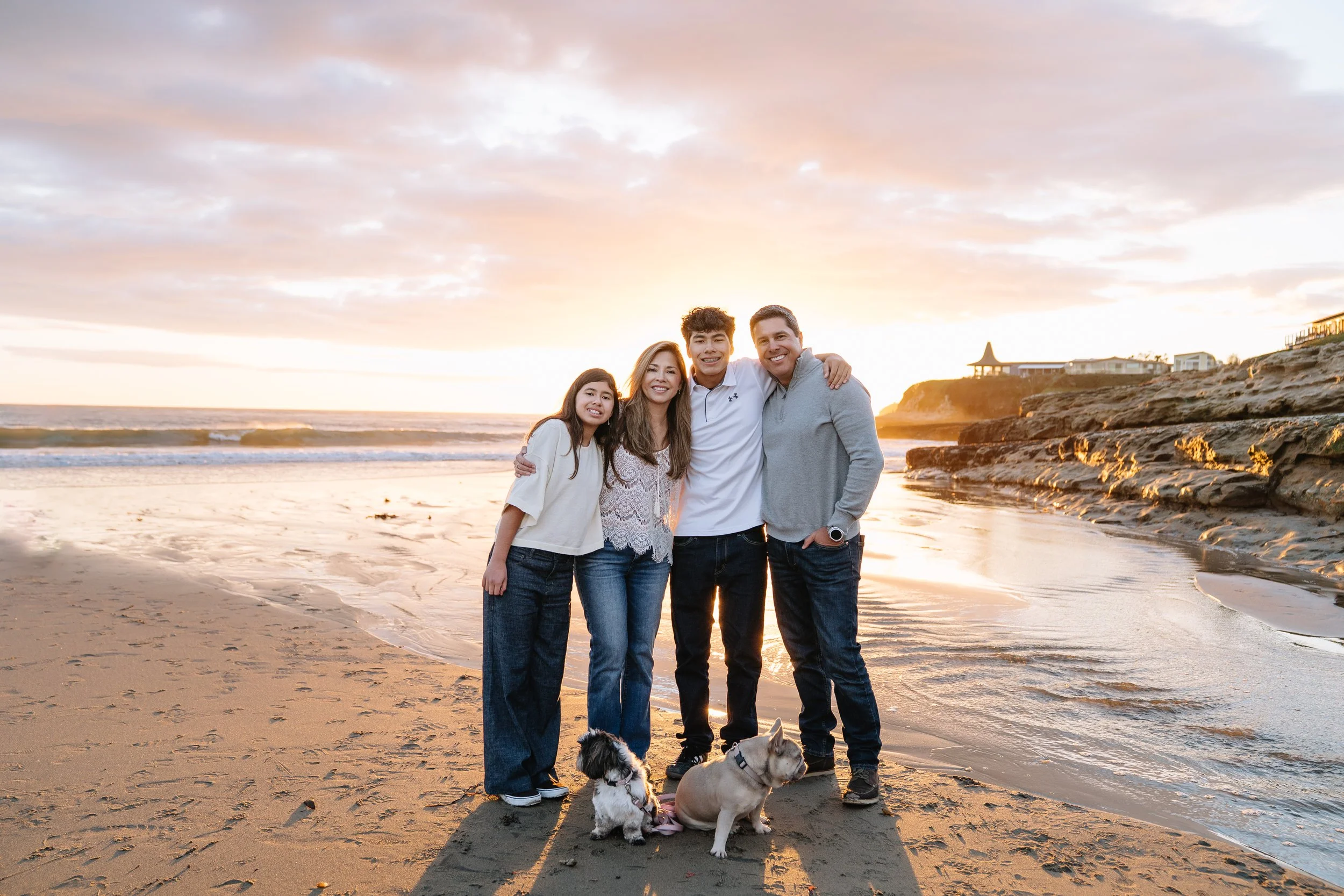 A family of four, along with two dogs, standing on a sandy beach at sunset with ocean waves and rocky cliffs in the background.