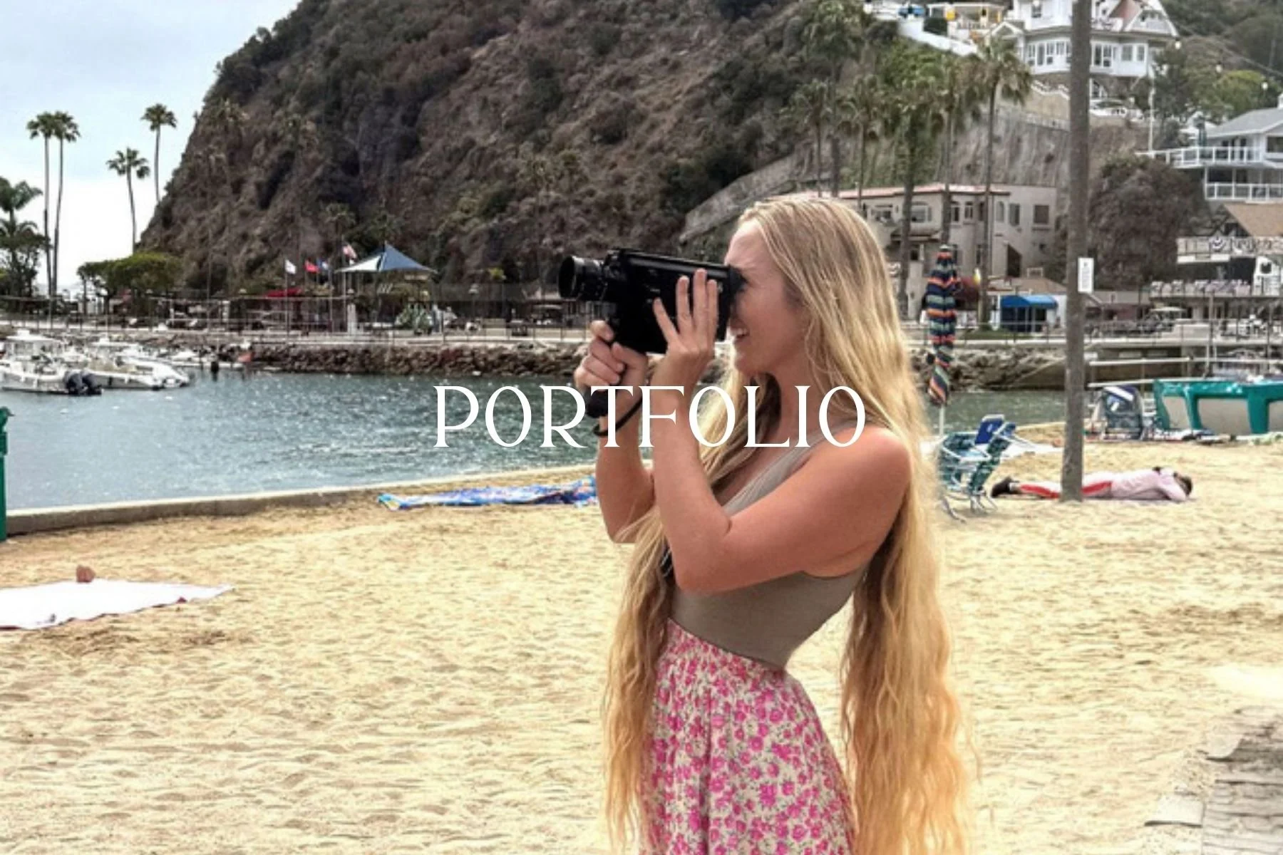 A young woman with long blonde hair, wearing a light-colored tank top and a pink floral skirt, takes a photograph with a vintage film camera at a beach. The background shows a rocky hillside with houses, palm trees, and a marina with boats.