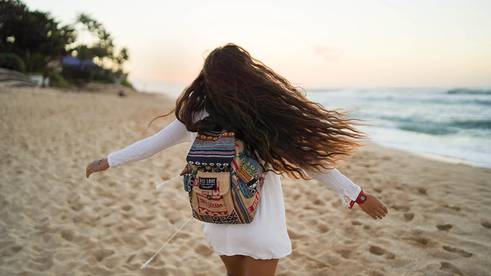 A woman with long, wavy hair walks on the beach at sunset, wearing a white dress and a colorful backpack.