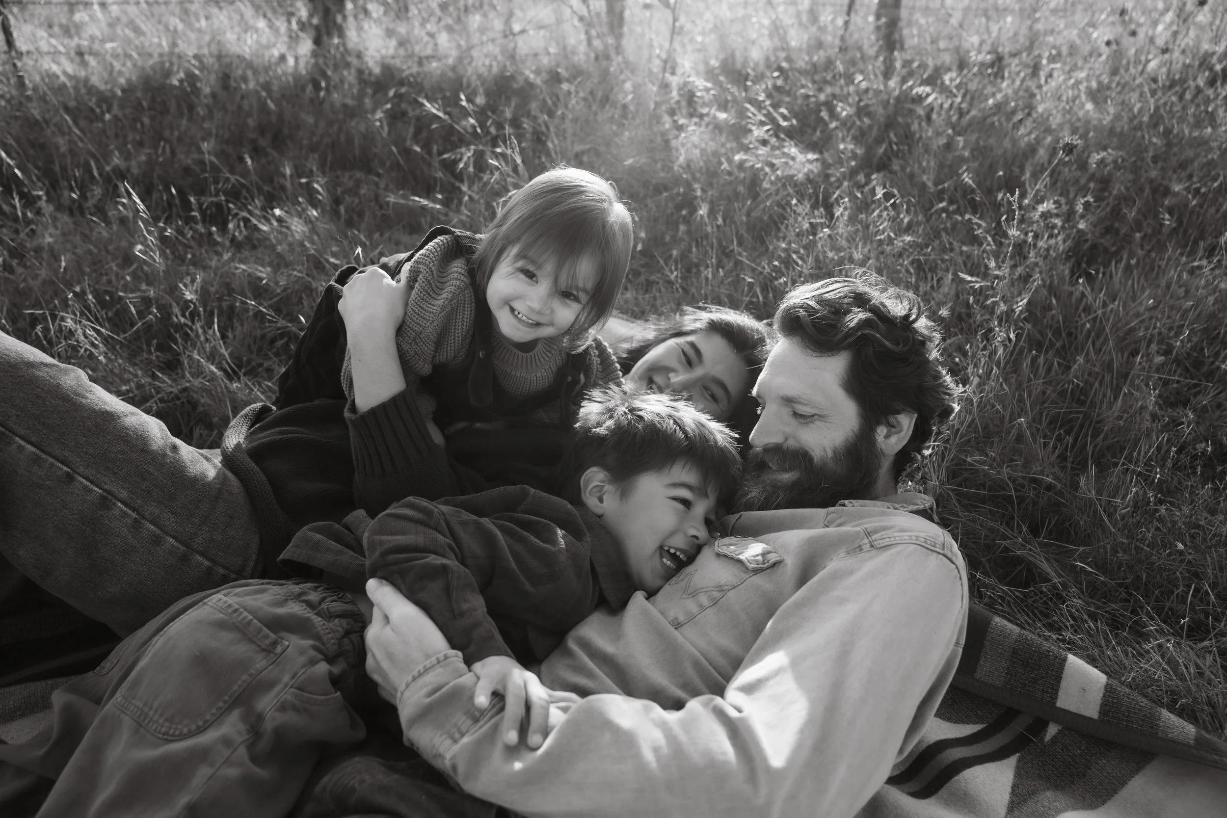 A family of four lying on the grass, smiling and playing together outdoors.