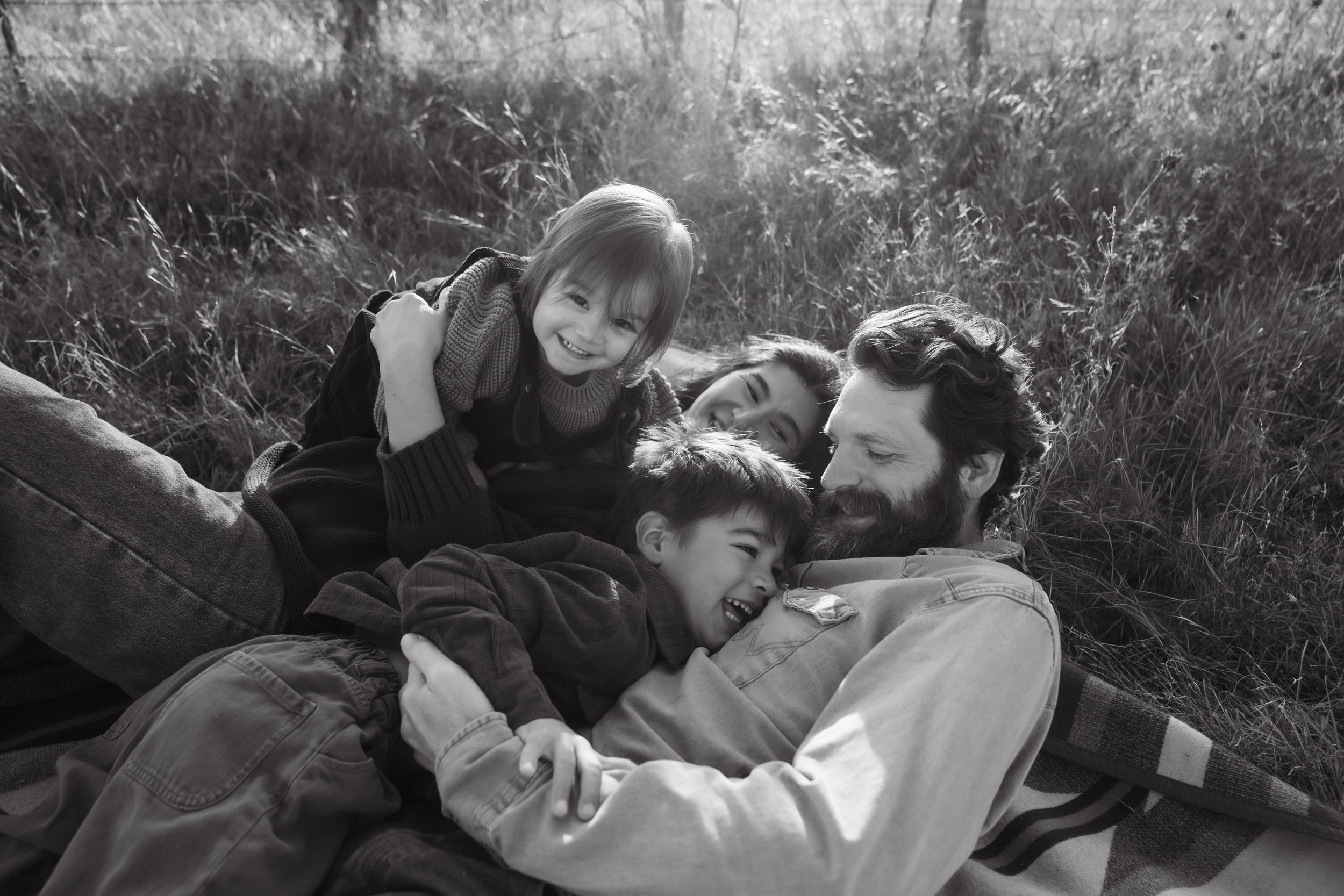 A family of four, including two children, lying on the grass and playing together outdoors, smiling and laughing.