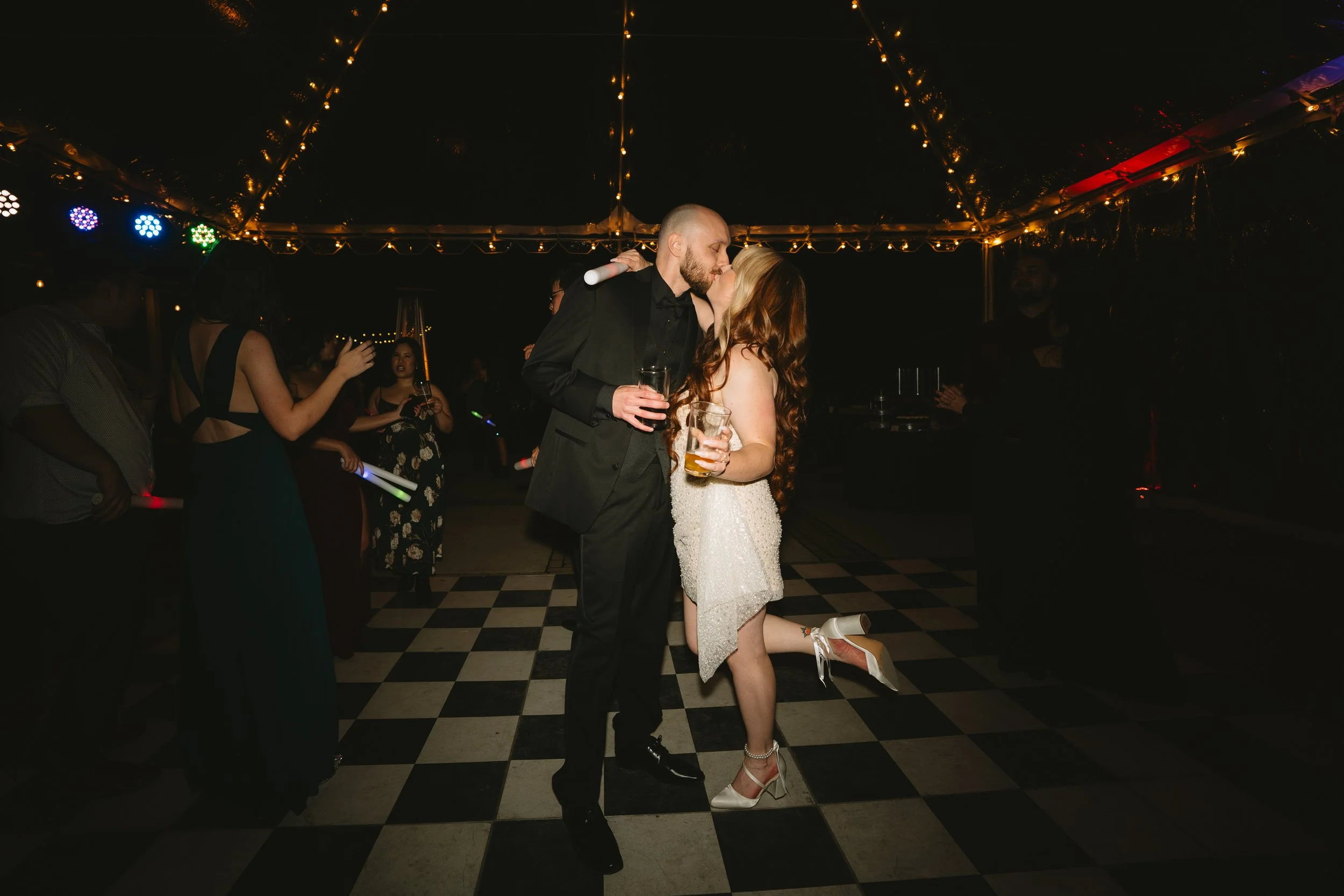 A couple kissing at a nighttime event or party under string lights, with the woman kneeling on a checkered dance floor and holding a drink, while other people are visible in the background.