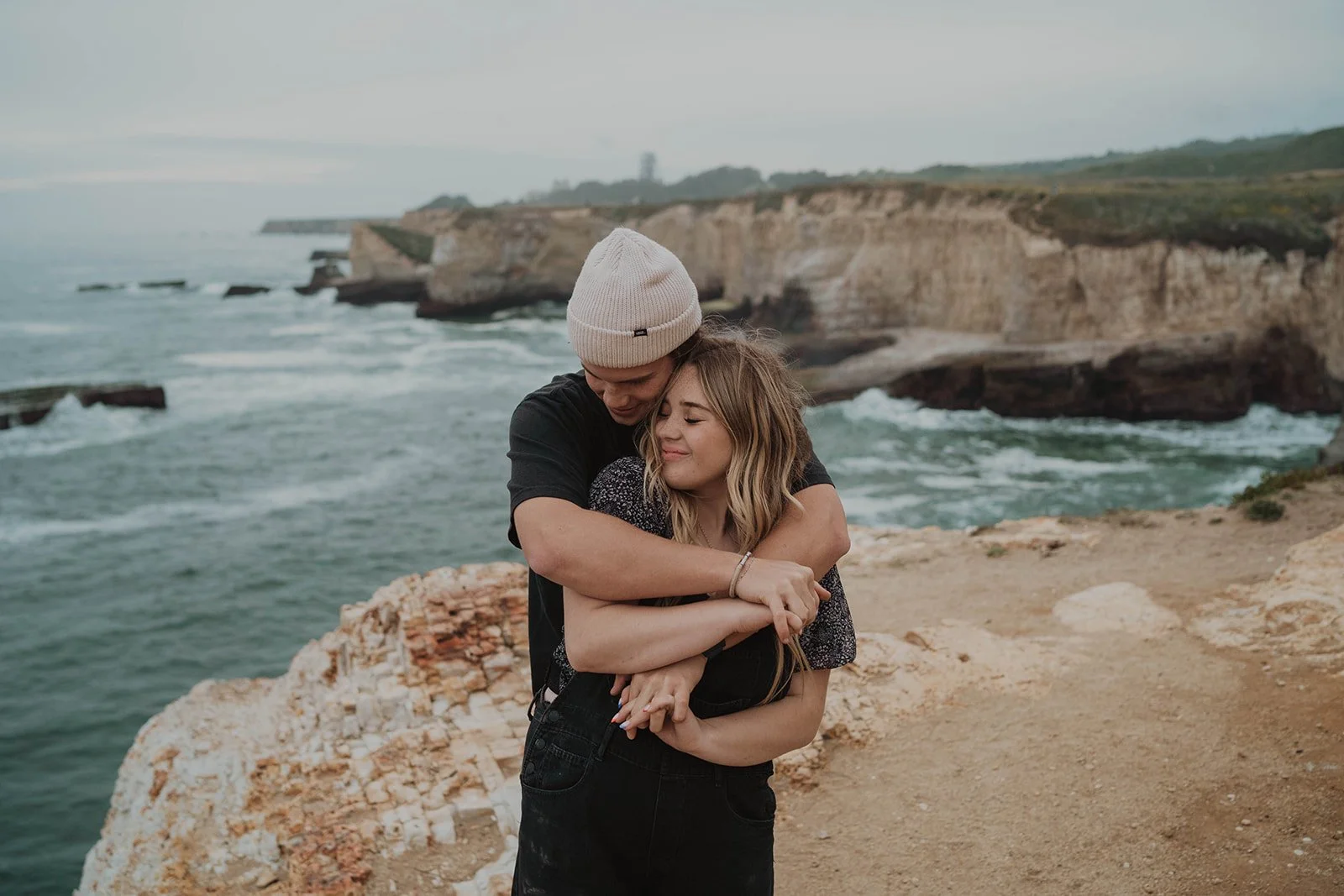 A young man and woman hugging on a cliffside near the ocean with cliffs and waves in the background.