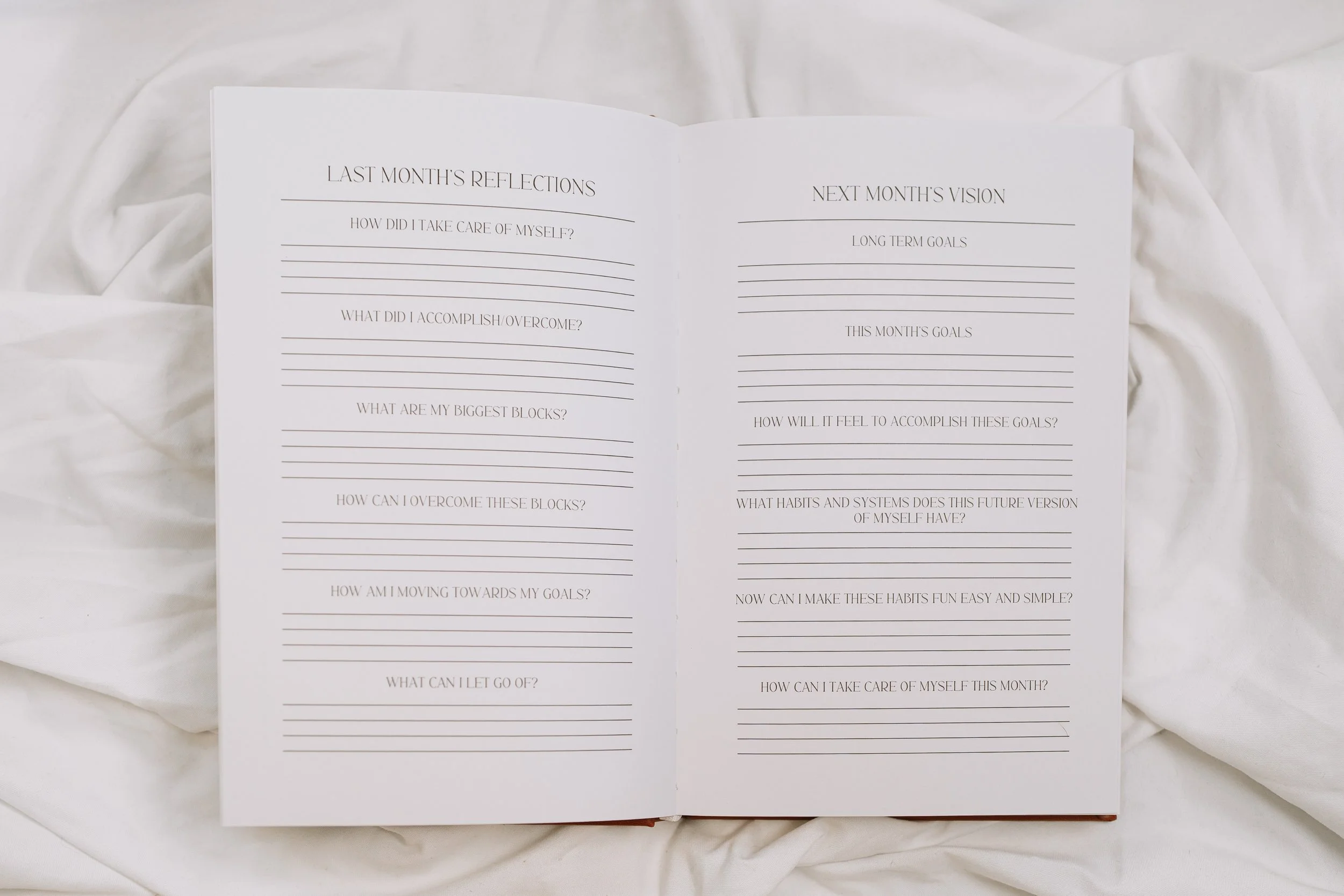 An open journal on a white fabric surface with pages containing prompts for reflections and goal setting for the past and upcoming month.