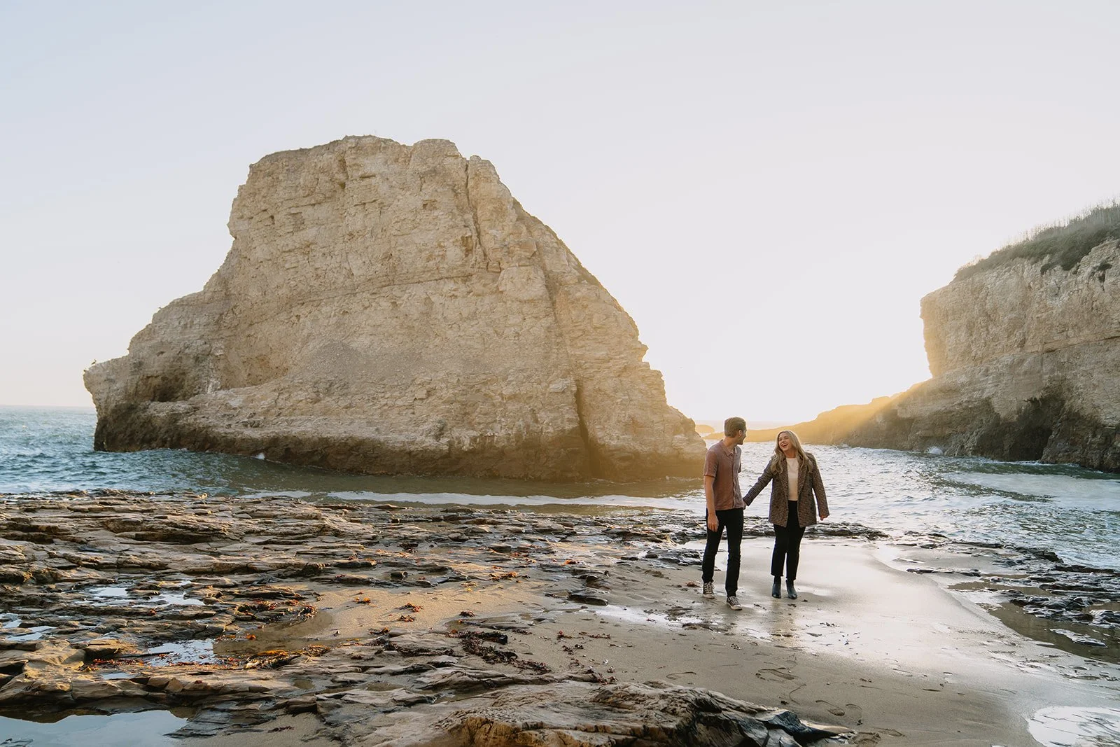A couple holding hands walking along a rocky beach with large cliffs and the ocean in the background during sunset.