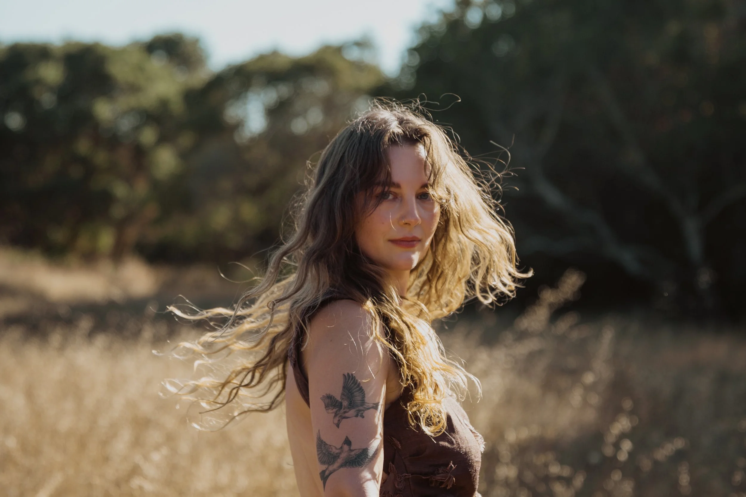 A woman with wavy hair and a tattoo of birds on her upper arm stands outdoors in a field with trees in the background, sunlight illuminating her face.