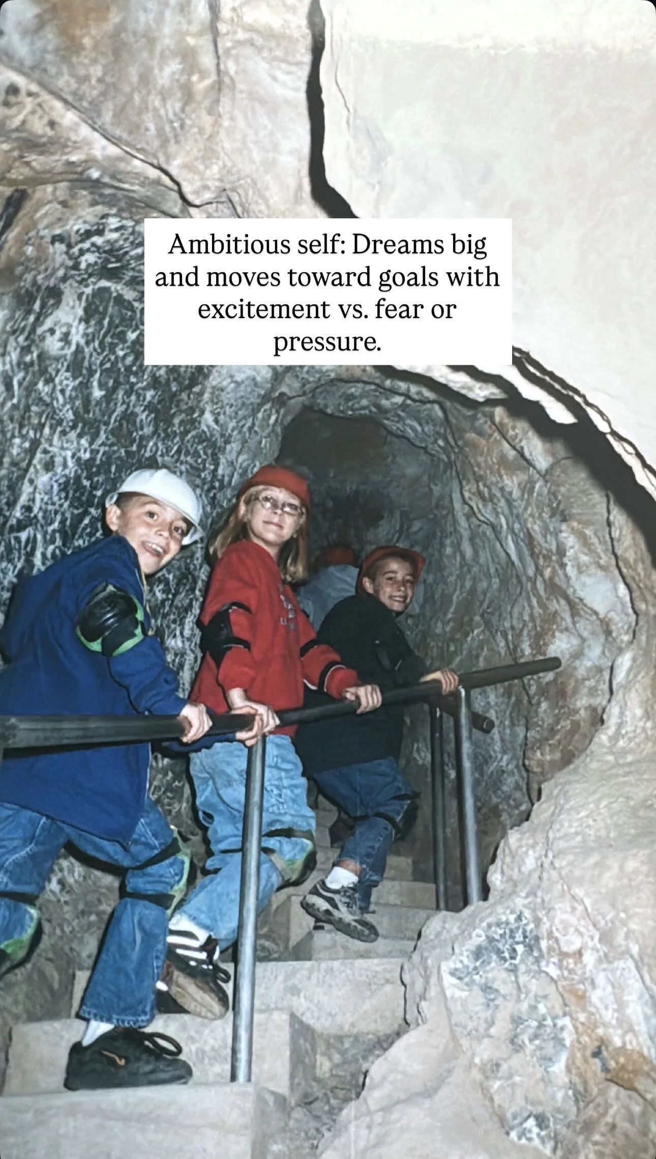 Three children inside a cave, standing on stairs and leaning on a metal railing, smiling at the camera.