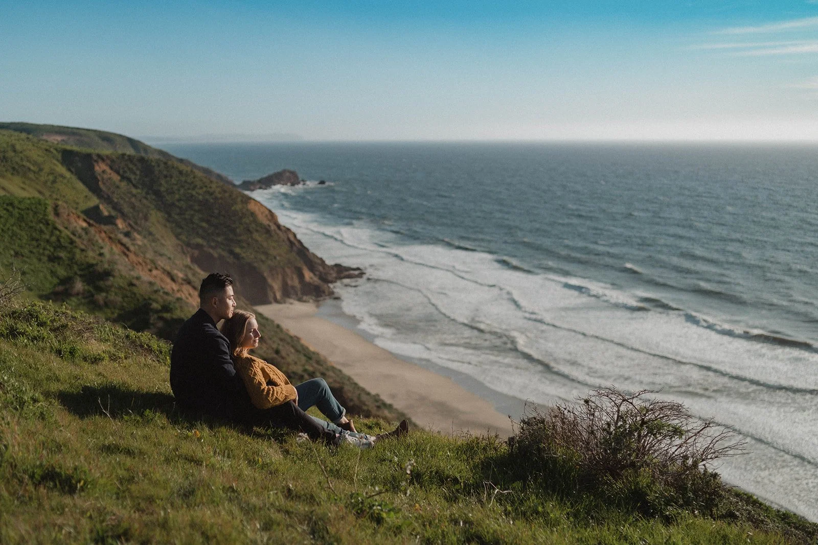 A couple sitting on a grassy hill overlooking the ocean and a sandy beach, with cliffs in the background on a clear day.
