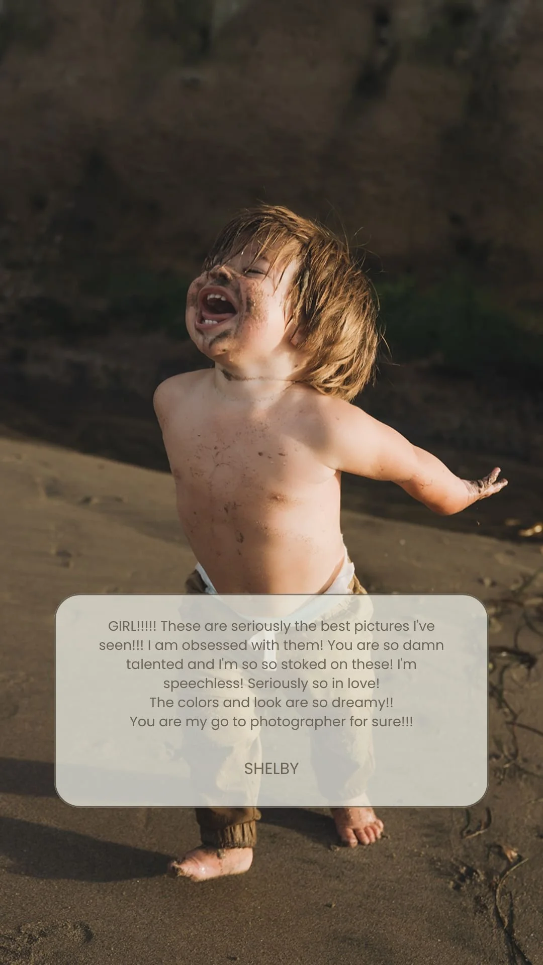 Young boy with messy hair, dirt on face and body, shirtless and wearing pants, standing on a sandy beach with a blurred rocky background, expressing joy or excitement.