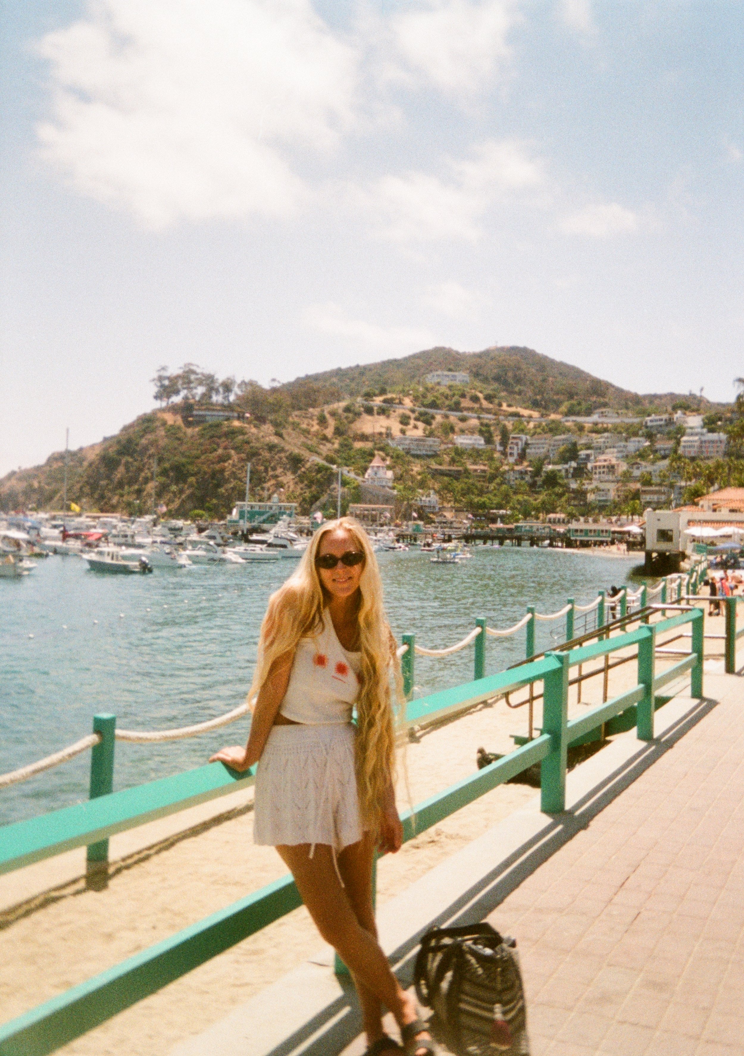 A woman with long blonde hair wearing sunglasses, a white t-shirt with a design, and a white skirt, standing on a waterfront promenade with boats in the water and a hillside with houses in the background.