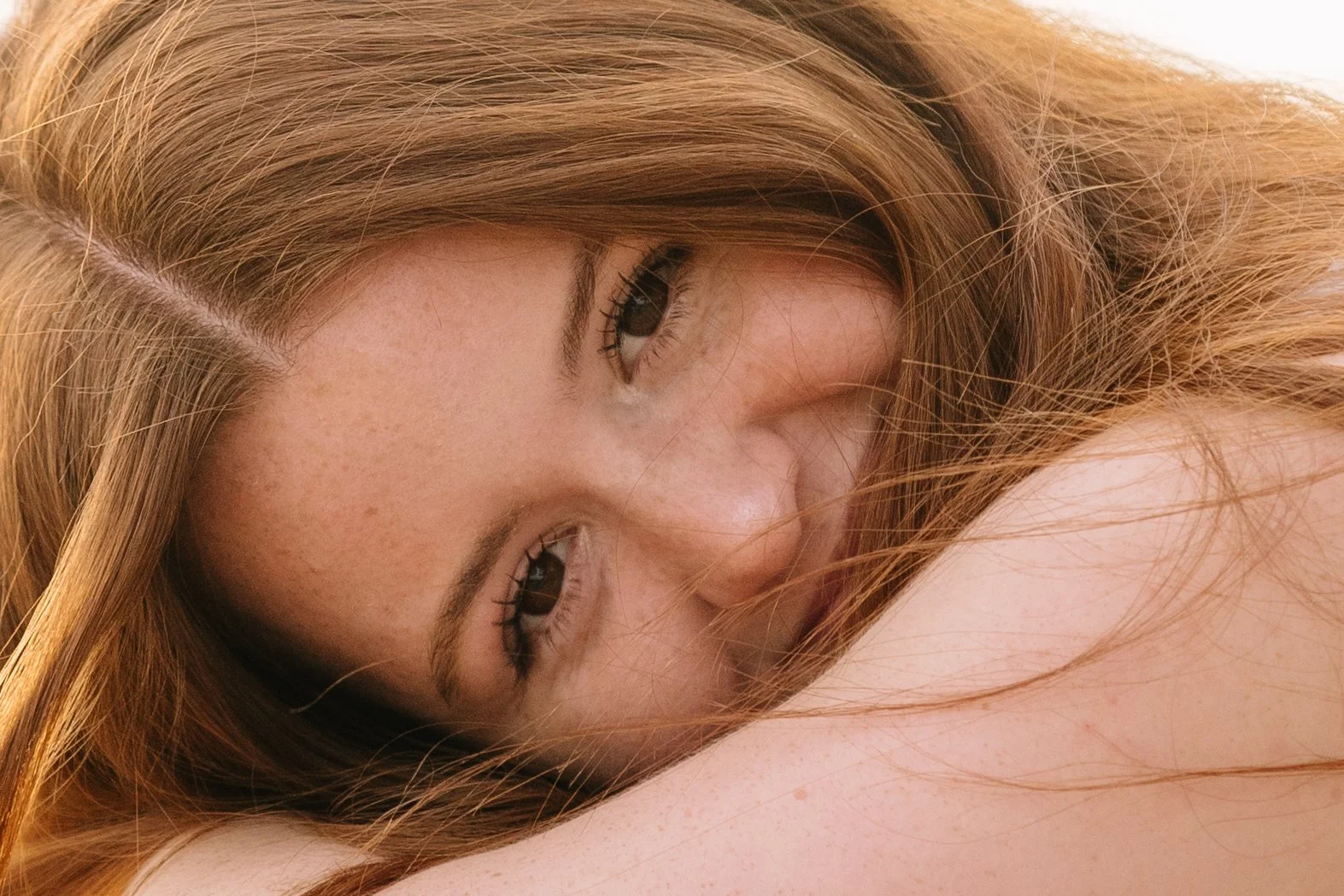 Close-up of a woman with ginger hair, lying down with her face resting on her arm, looking directly at the camera with a soft expression.