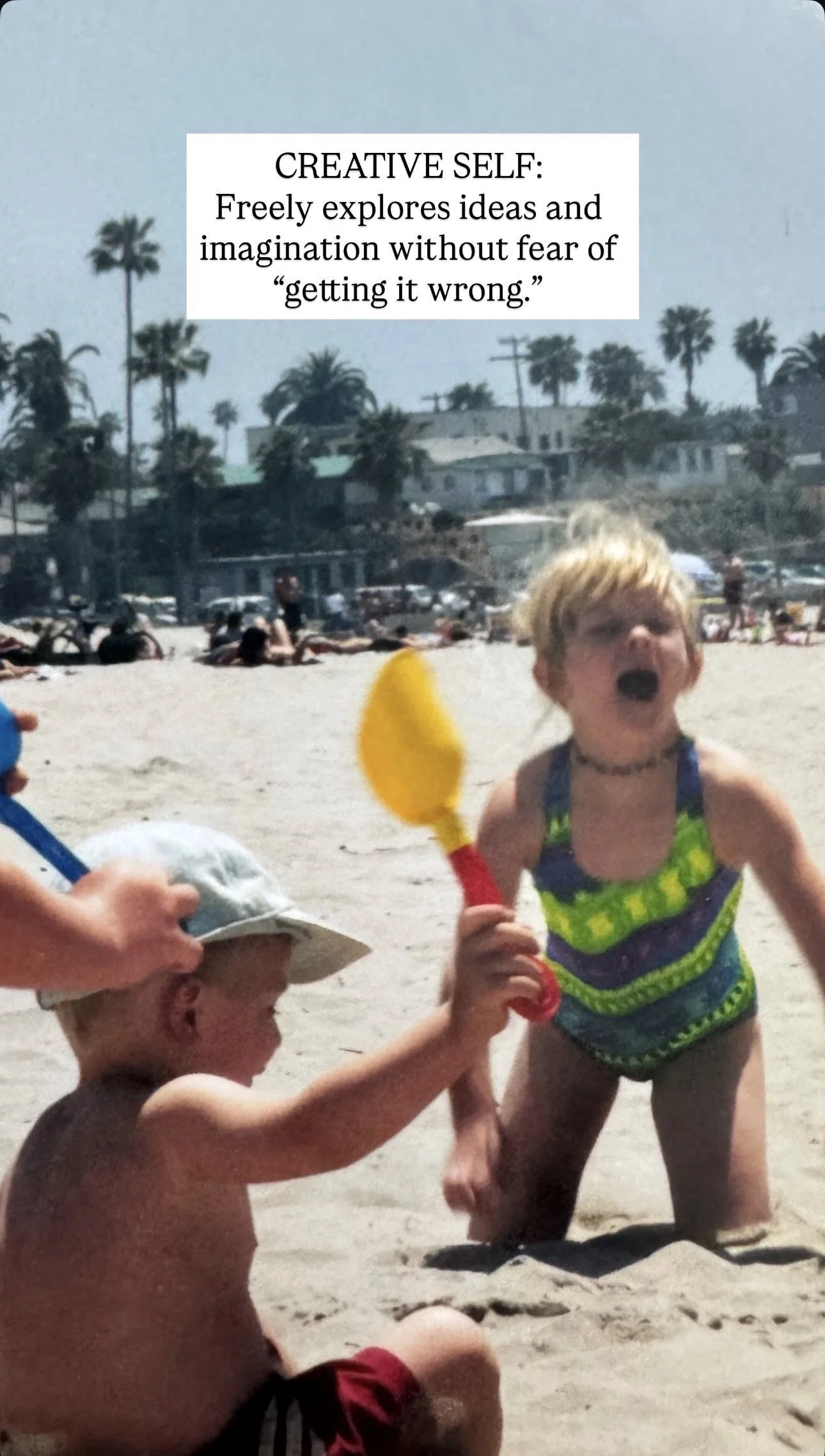Two young children playing with a yellow toy shovel on a sunny beach, with palm trees and houses in the background.