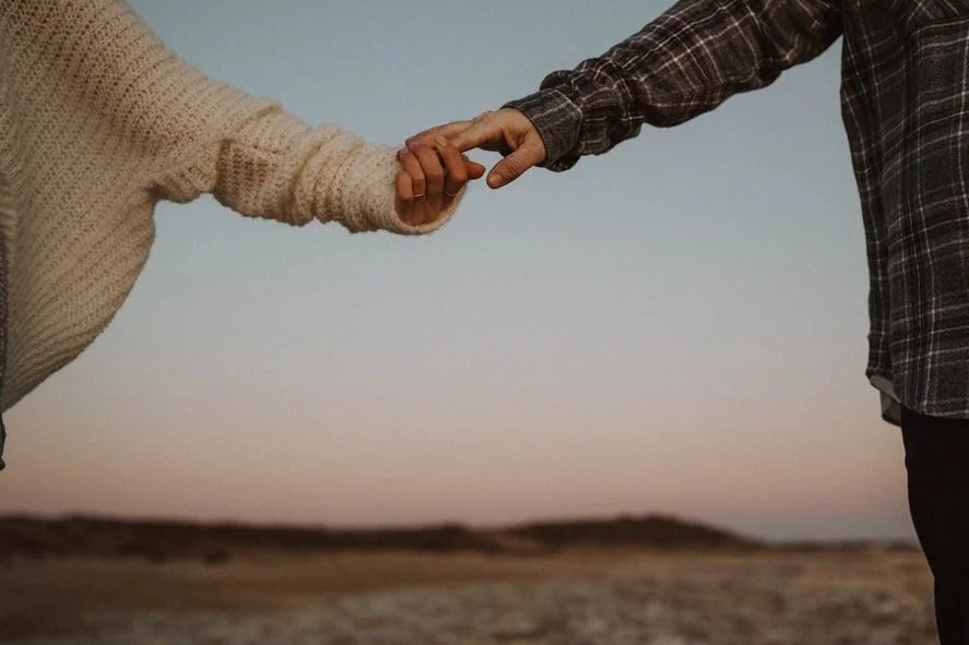 Two people holding hands outdoors during sunset or dusk, with a landscape in the background.
