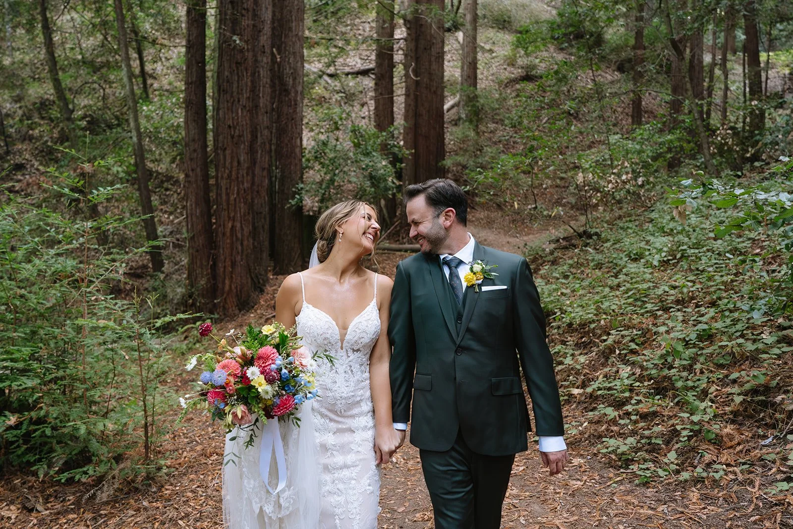 A bride and groom holding hands and smiling at each other in a forested area during their wedding.