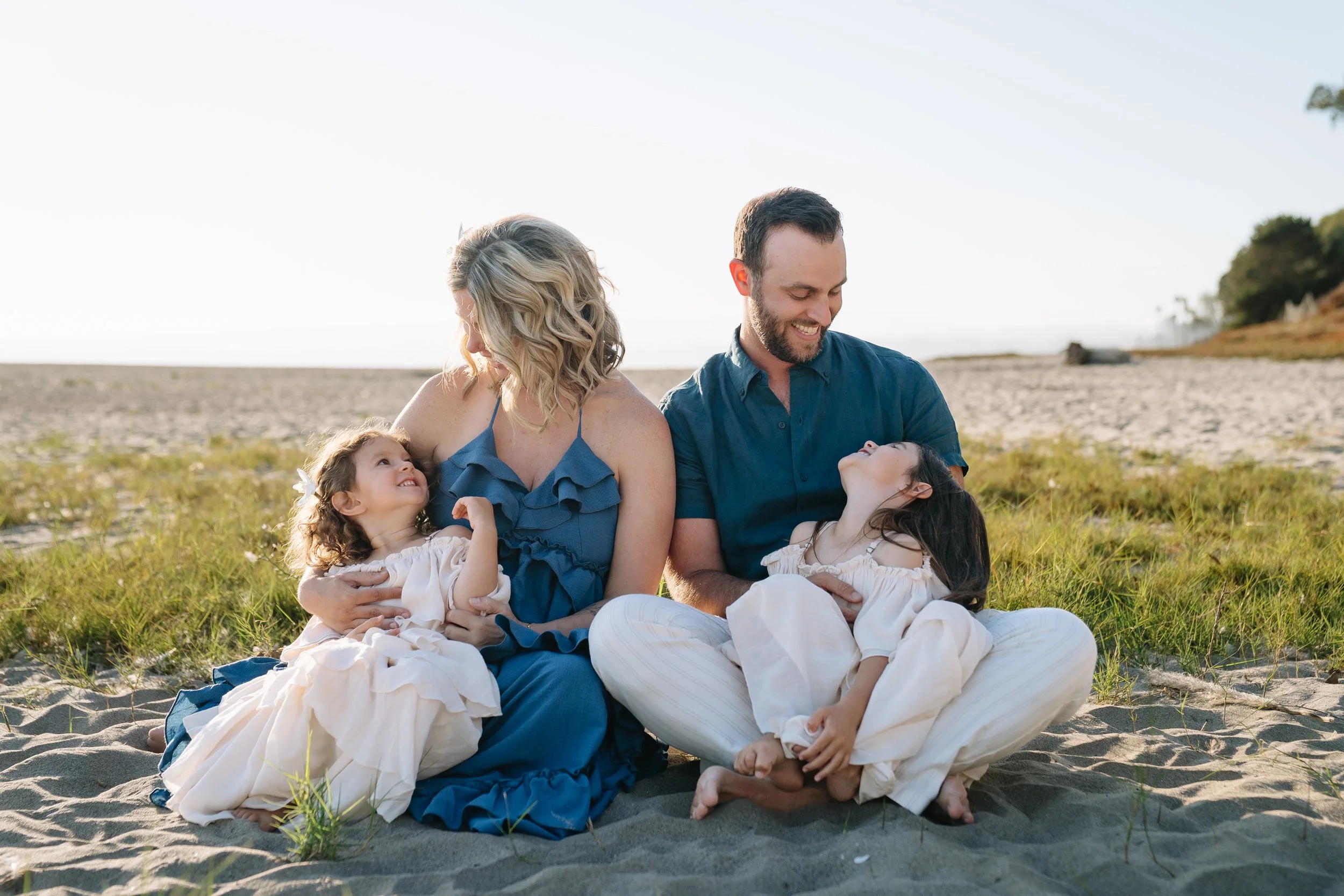 A happy family of four sitting on the beach, two parents and two young girls, smiling and looking at each other, with grass and sand in the background.