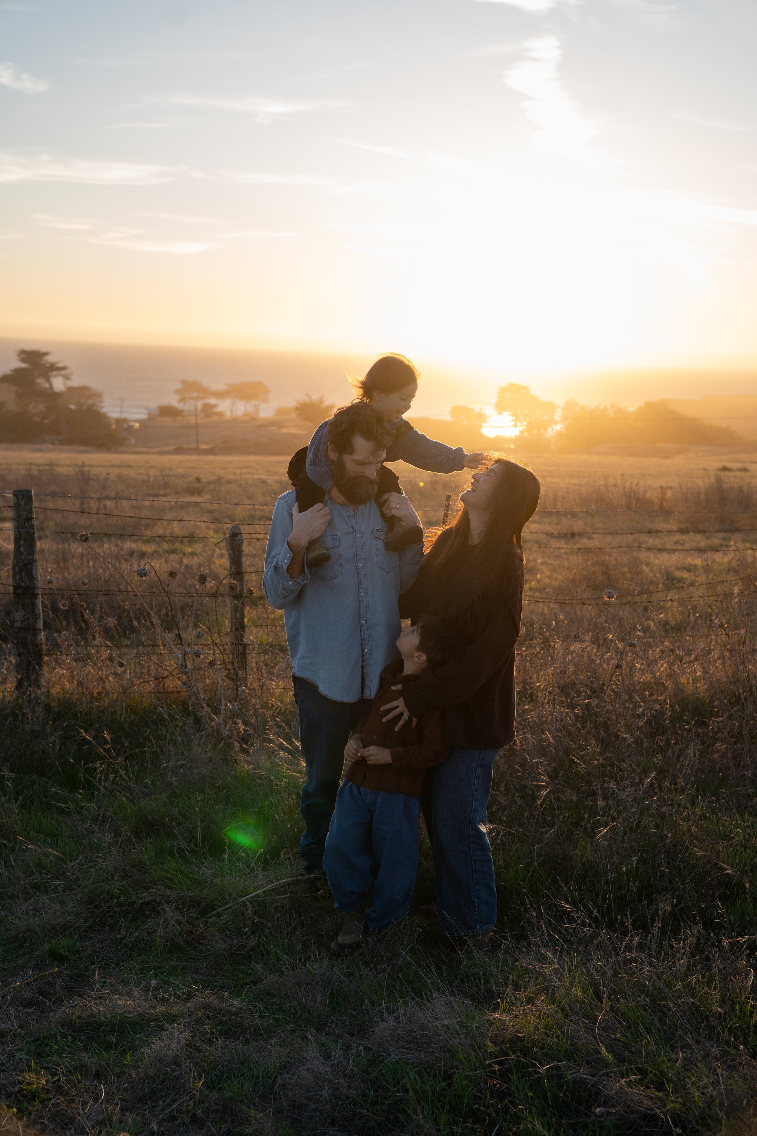 A family of four, including two adults and two children, enjoying a sunset outdoors near a fence, with trees and the ocean in the background.