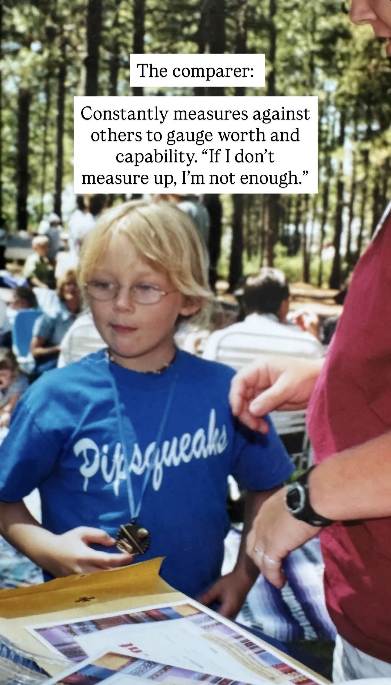 A boy with blond hair, glasses, wearing a blue 'Disquakes' T-shirt, stands outdoors in a wooded area during a gathering, holding a medal and looking to the side, with people sitting at tables in the background.