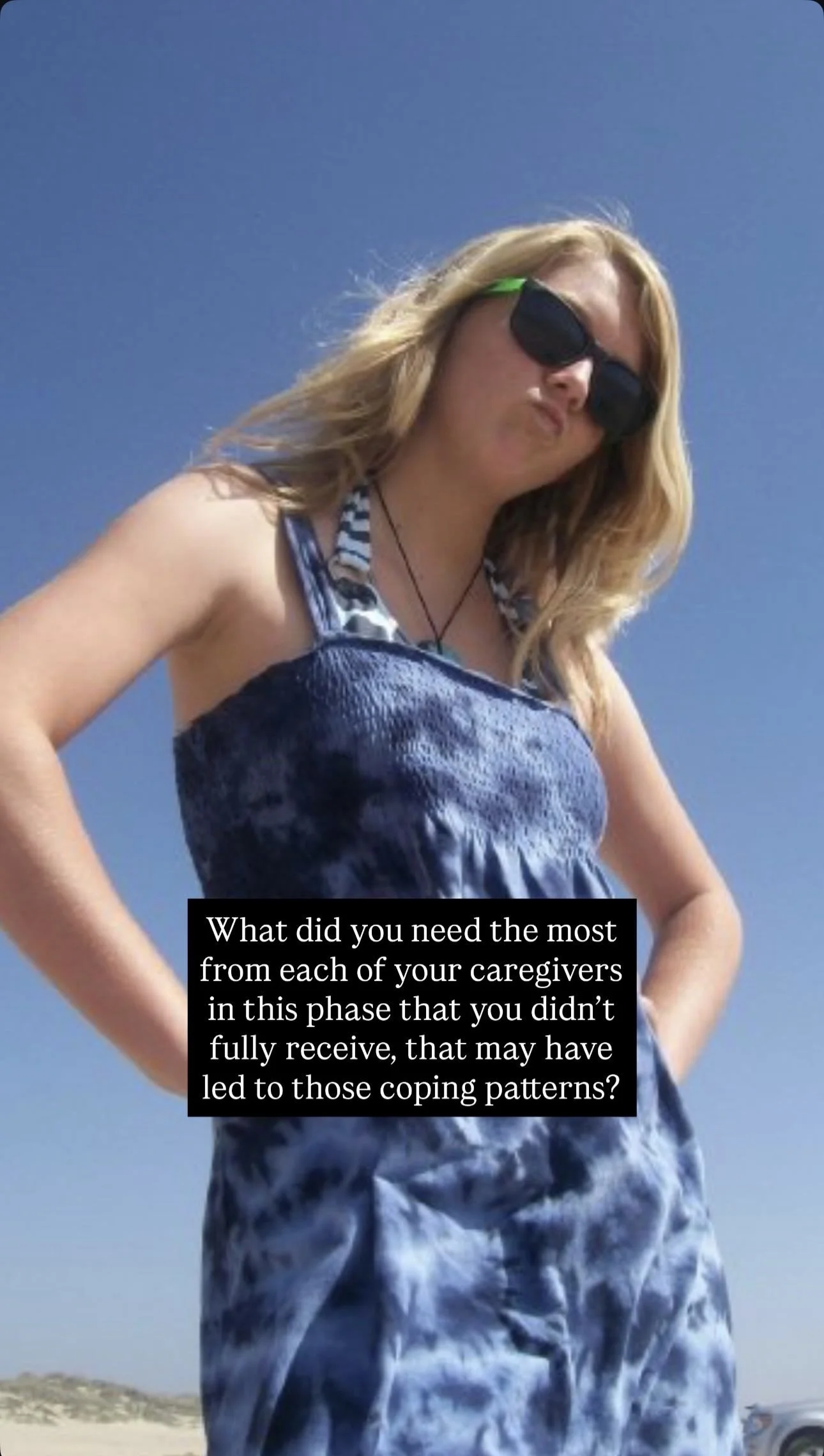 A woman with blonde hair wearing black sunglasses, a blue tie-dye dress, and a striped swimsuit top, standing outdoors against a clear blue sky.