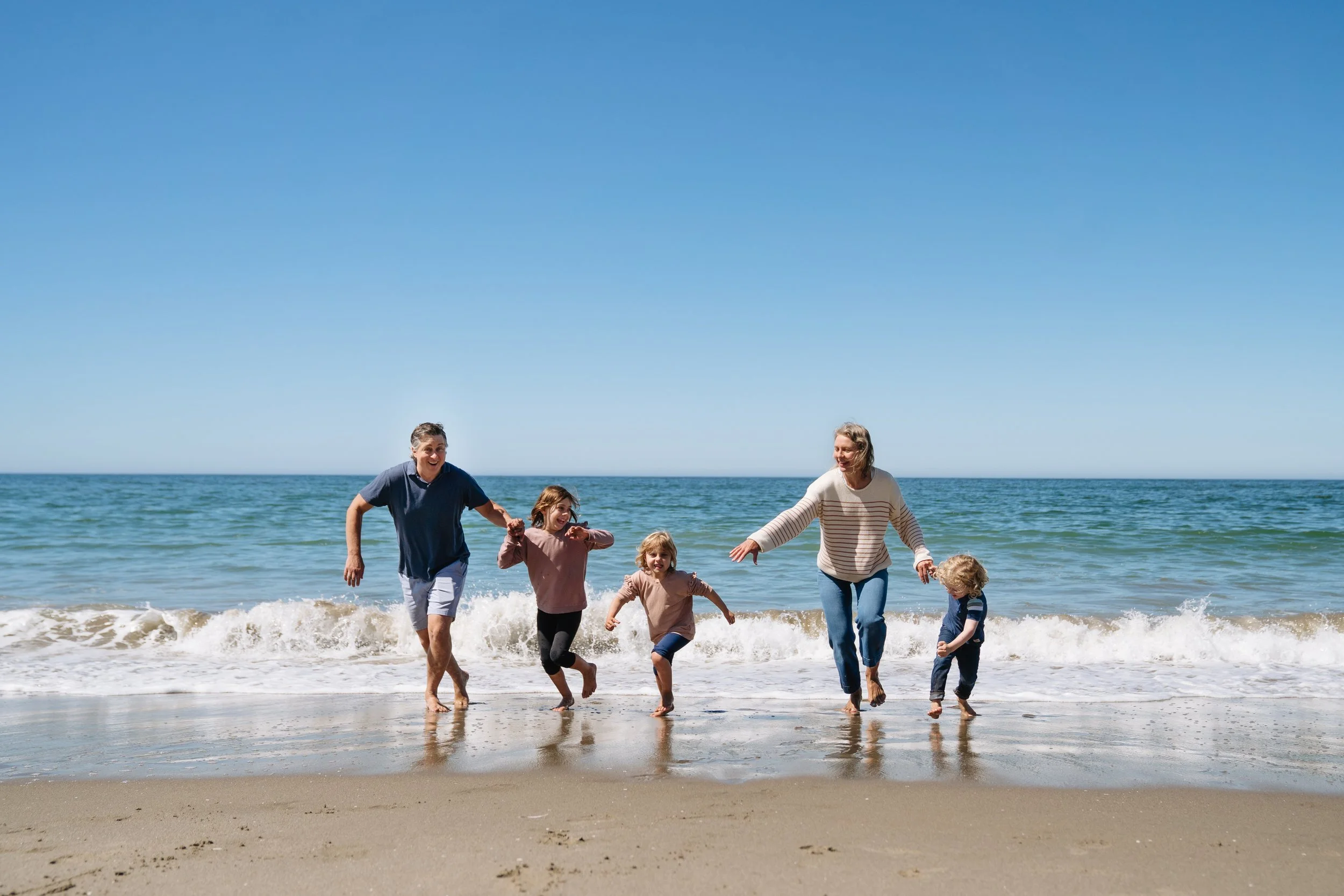 Family of six playing and running on the beach near the ocean with blue sky.