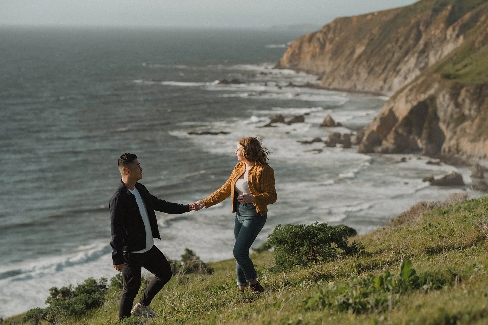 A woman and a man holding hands on a grassy hillside overlooking the ocean with cliffs in the background.